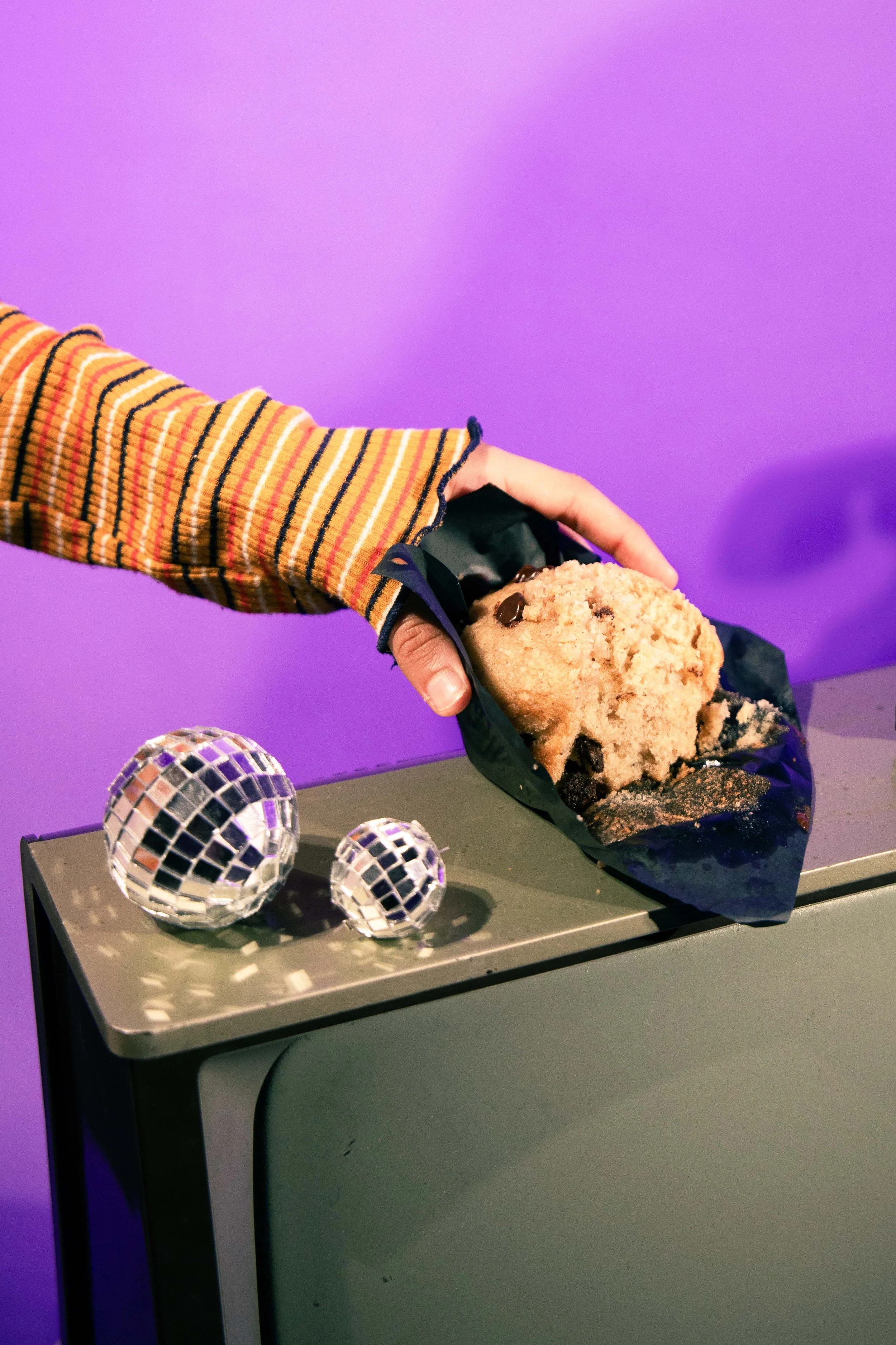 A hand in a striped long sleeve shirt holding a chocolate chip cookie wrapped in black paper, with three disco ball ornaments on a gray surface and a purple background.