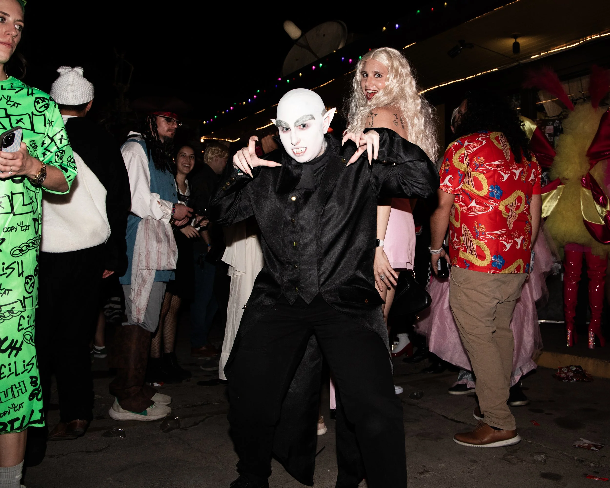 Two people in costume, one with white face and pointy ears dressed as a vampire and a woman with curly blonde hair posing together at a crowded party with colorful lights and costumes.