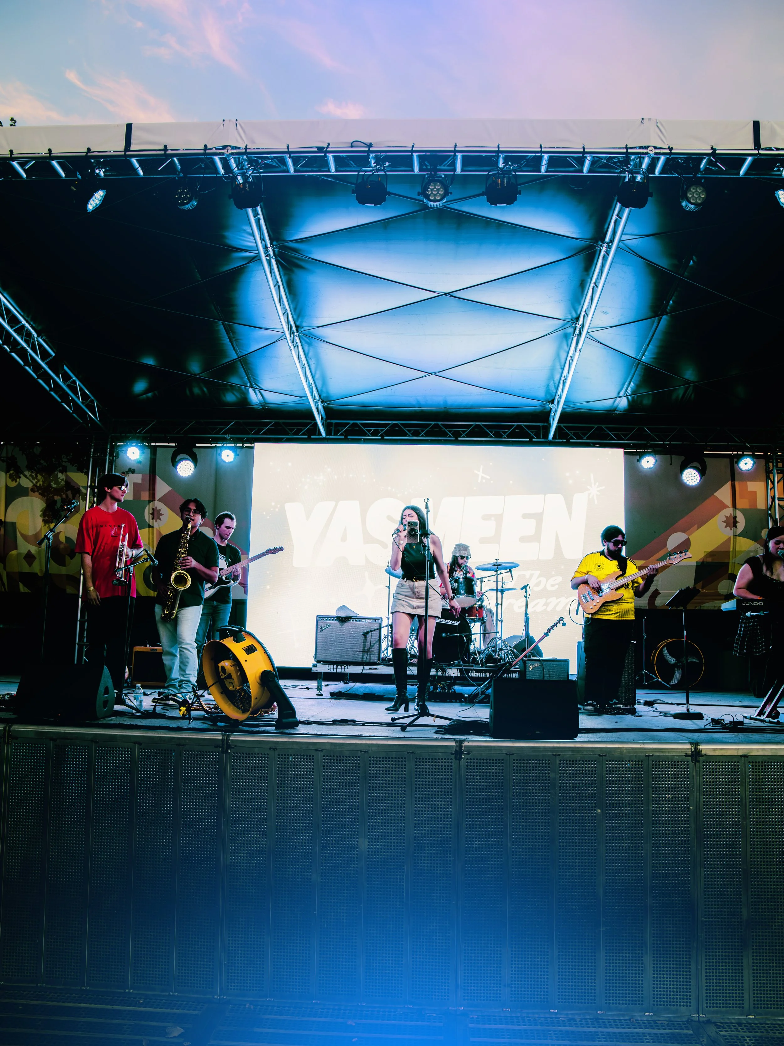 A band plays on an outdoor stage under a large black canopy, with a cloudy sky in the background. The band members include a woman singing at a microphone, a guitarist, a bassist, a drummer, and a saxophonist. Stage lights and equipment are visible.