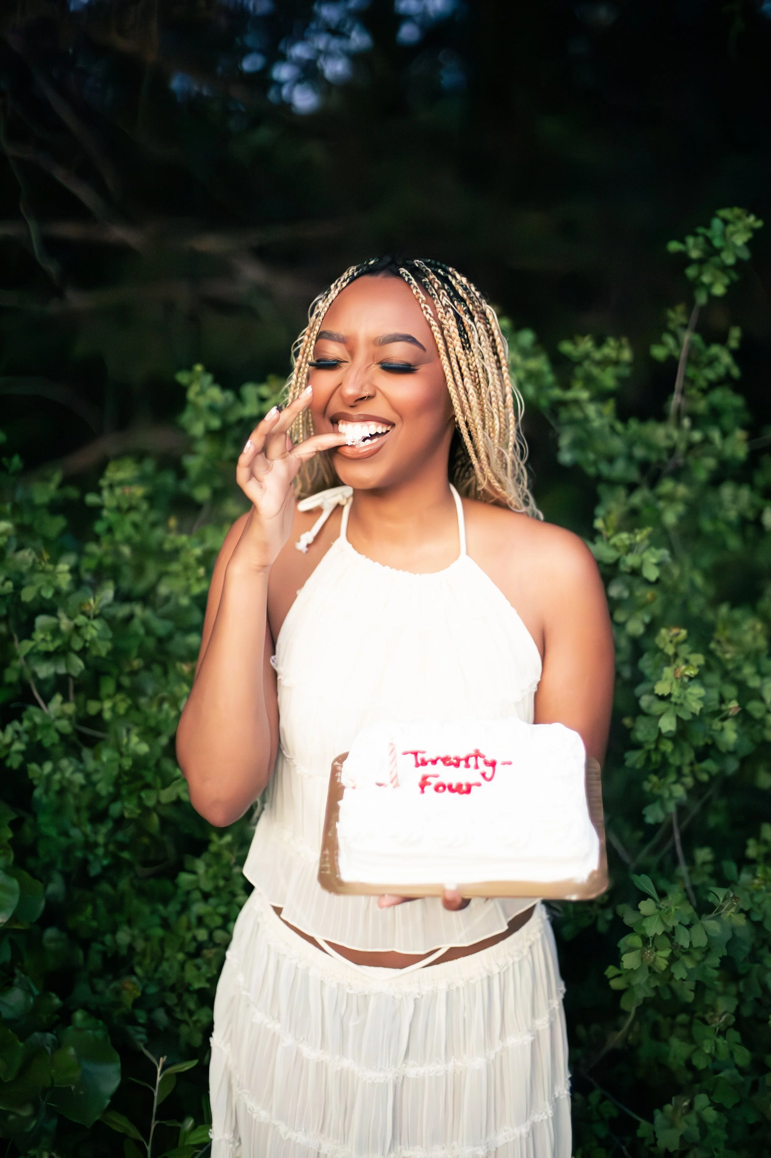 A woman with blonde braids wearing a white dress, smiling with eyes closed, holding a birthday cake that says 'Twenty-Four' in red text, outdoors with green foliage in the background.