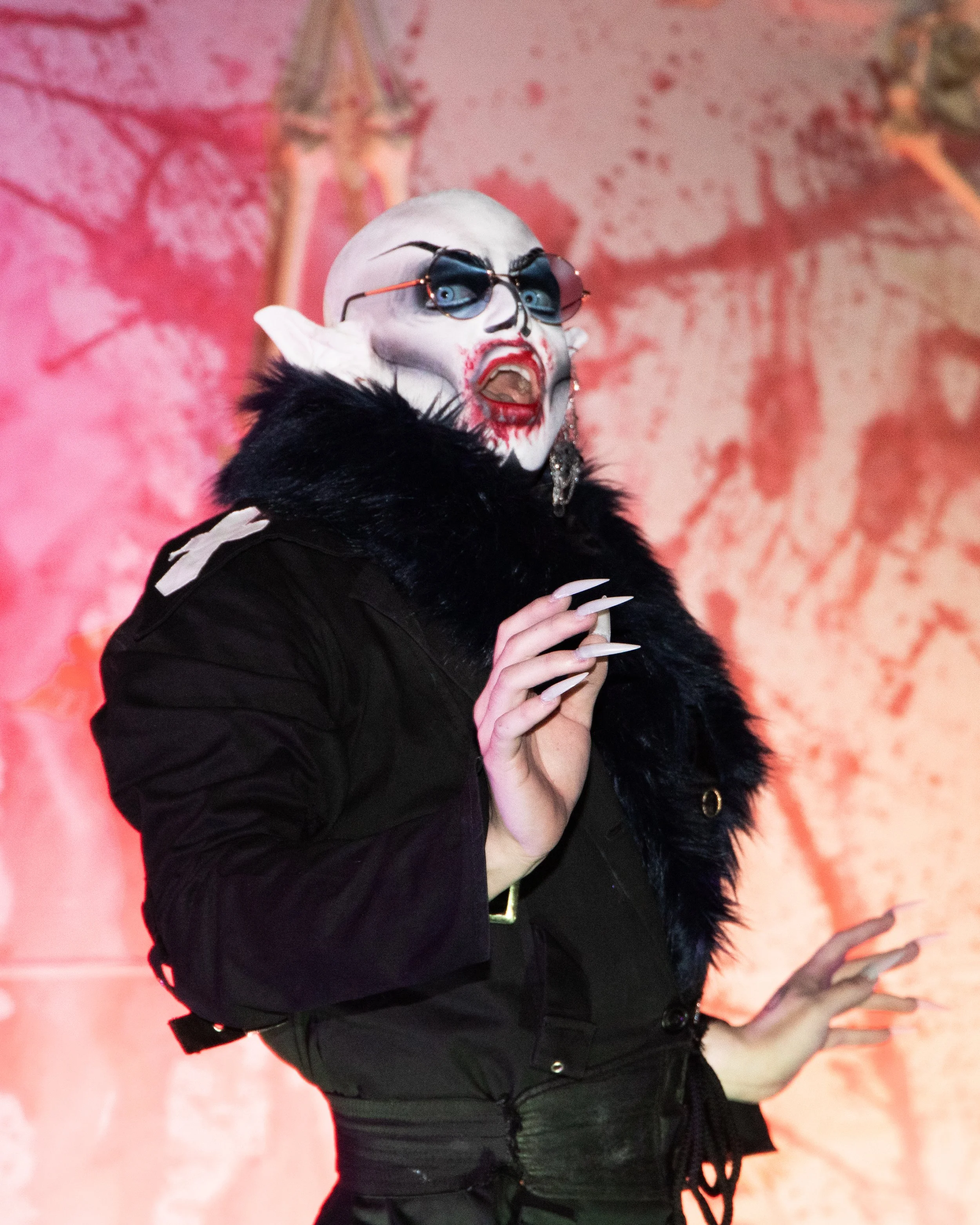 A person with face paint resembling a skull wearing glasses with blue lenses, black clothing with a fur collar, and long pointed nails, standing with arms crossed in front of a pink and orange wall with abstract patterns.