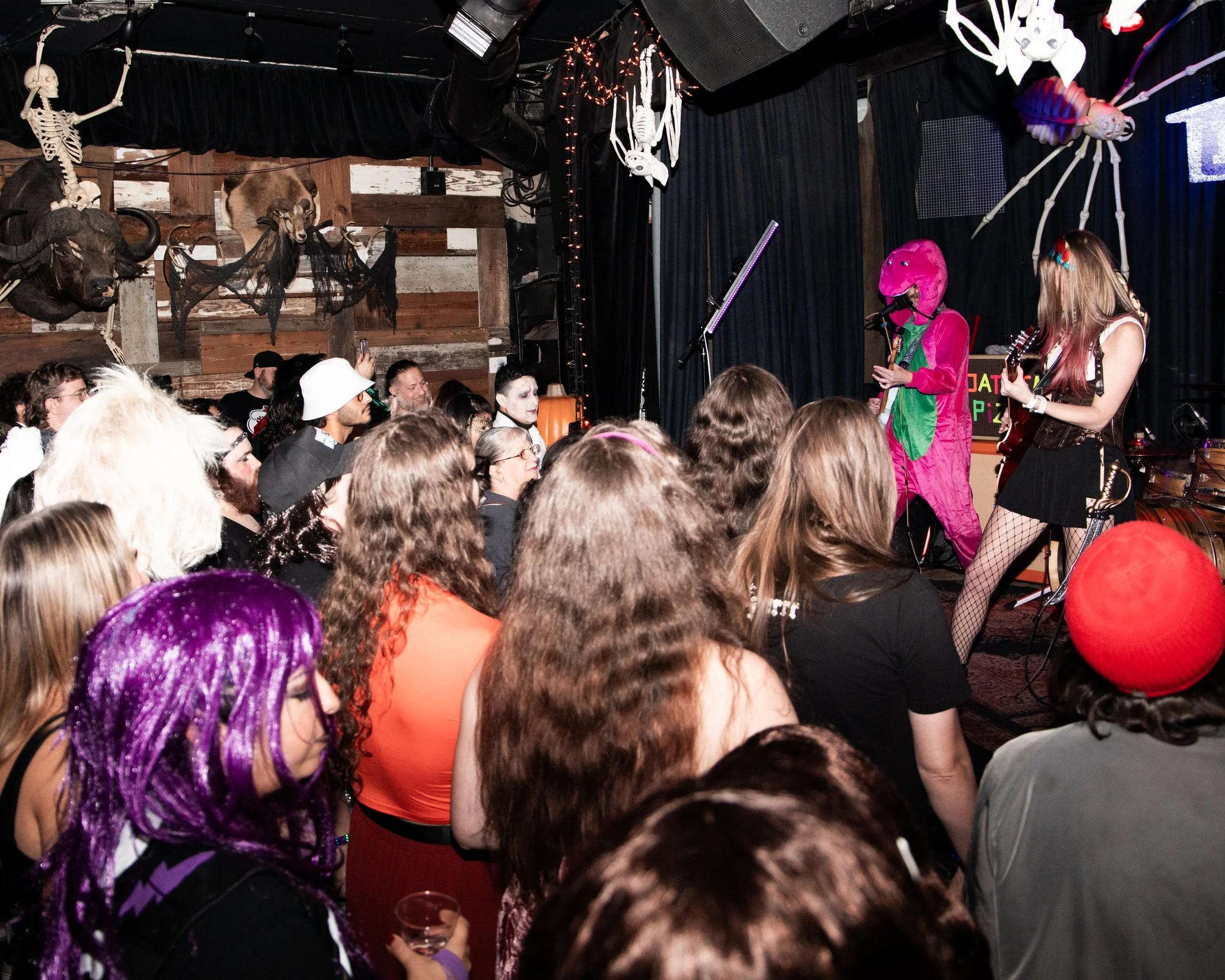 A crowded indoor event with people facing a stage where two performers are dressed in colorful costumes, including one in a bright pink dinosaur mask, and surrounded by hanging Halloween decorations such as skeletons, spiders, and other spooky decora