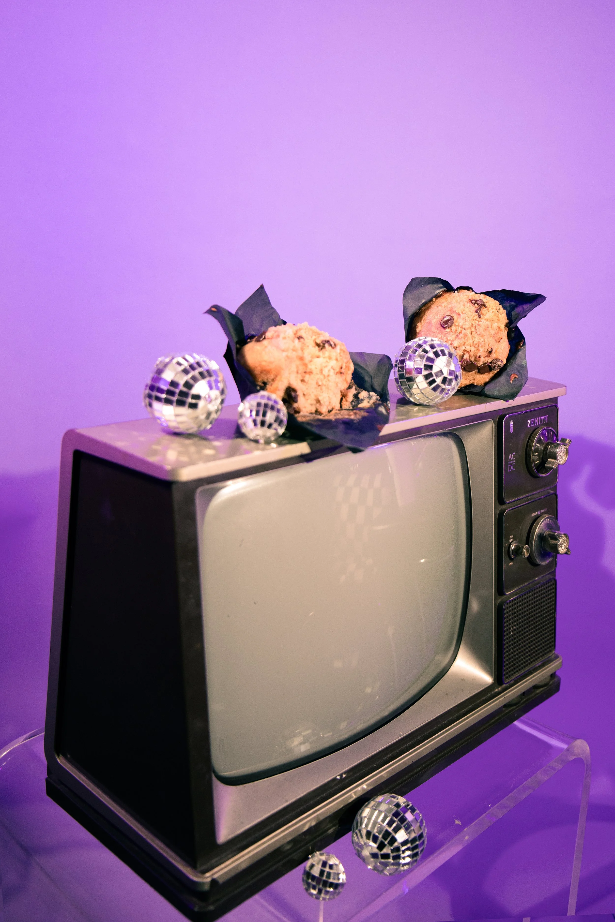 A retro-style microwave oven with a black and gray finish, topped with two chocolate chip cookies wrapped in black paper, and adorned with three small disco ball decorations, set against a purple background.