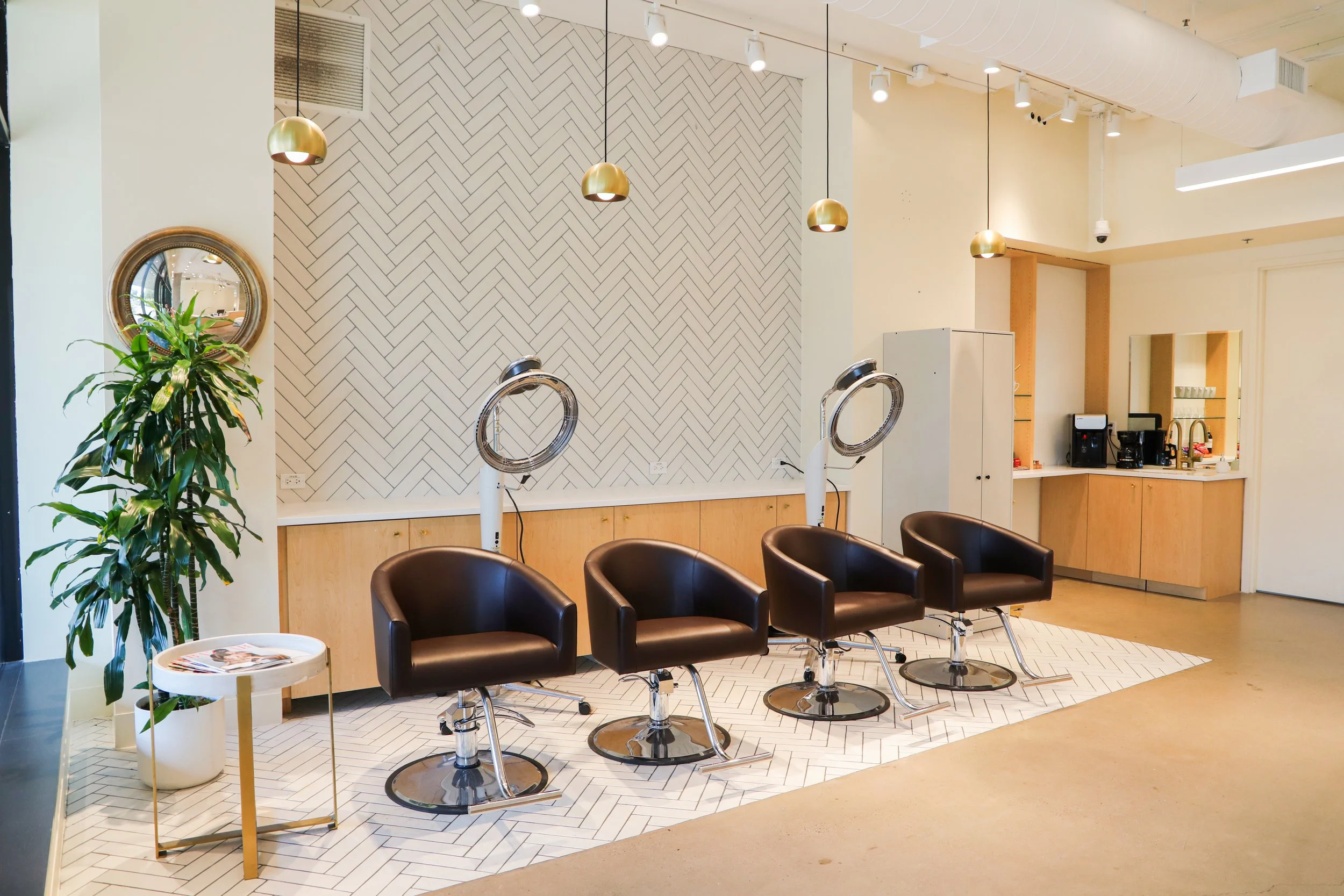 An interior view of a salon with five black styling chairs arranged in a row, a mirror, and a plant in a white pot. The walls are decorated with a herringbone patterned design and hanging pendant lights.