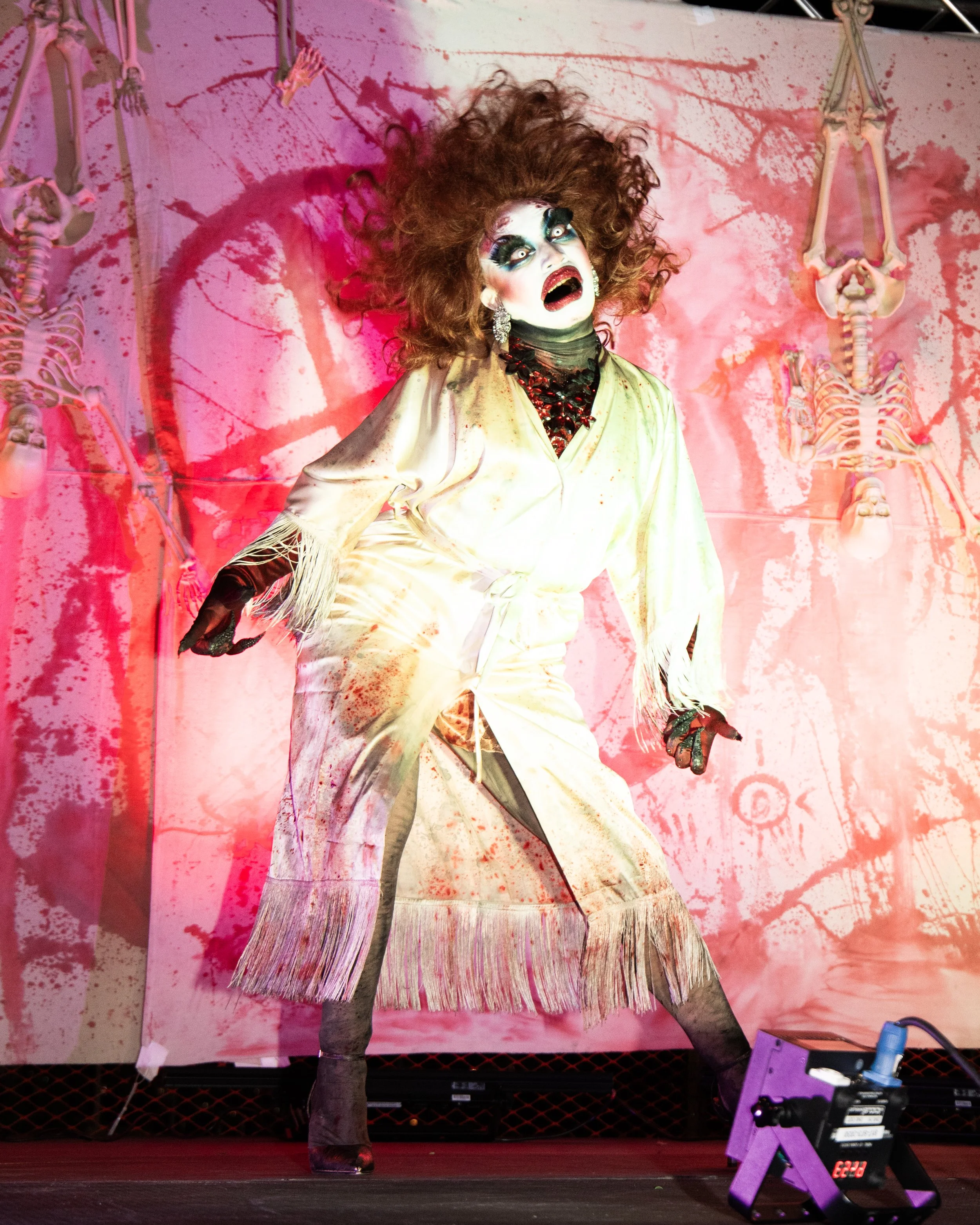 A person dressed in a theatrical costume with wild, curly hair and dramatic makeup, standing on a stage with skeleton decorations and splattered pink and red paint on the backdrop.