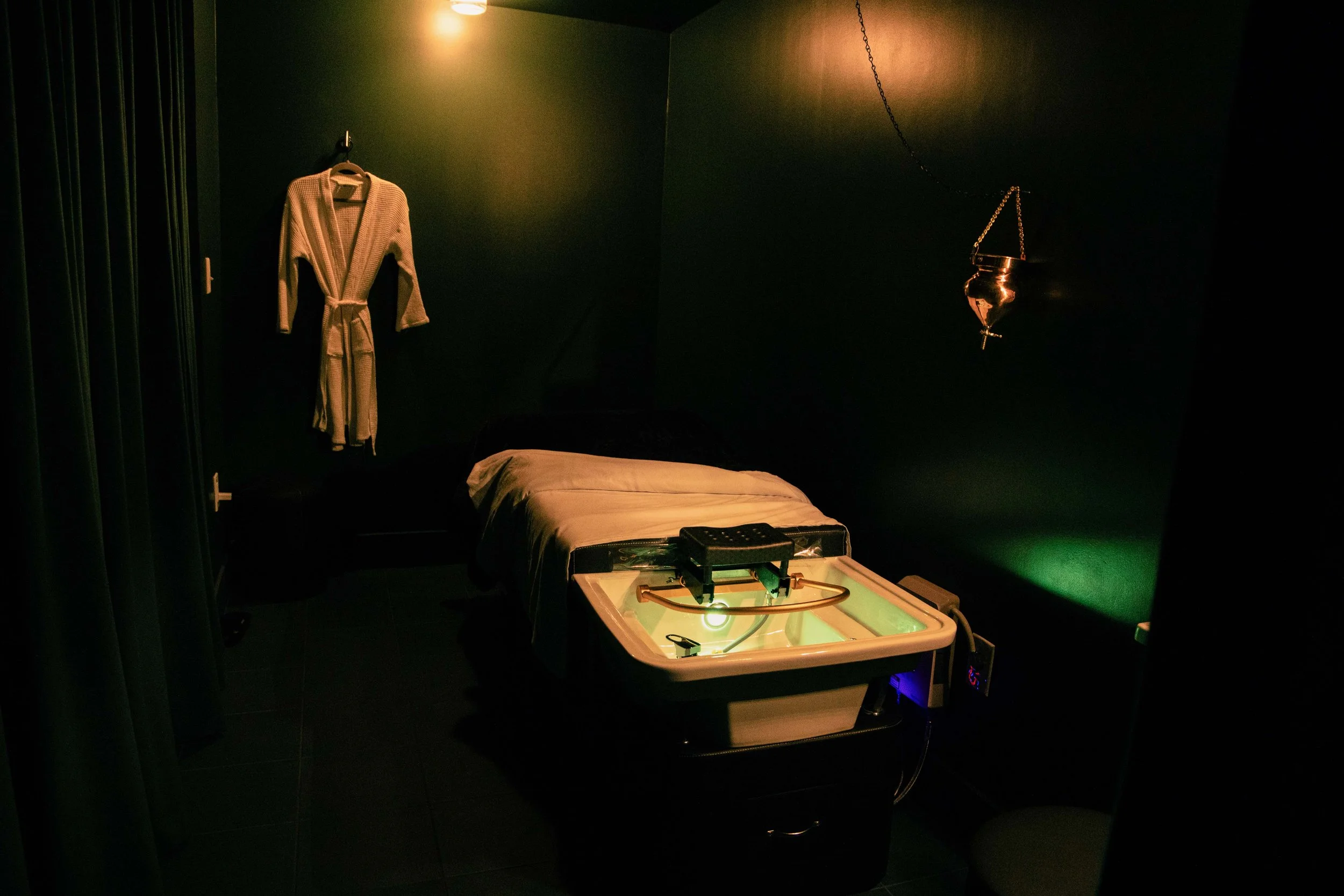 A dark room with a bed, a hanging metal incense holder, a robe hanging on the wall, and a light illuminating the space, with a small table and a game or device on it.