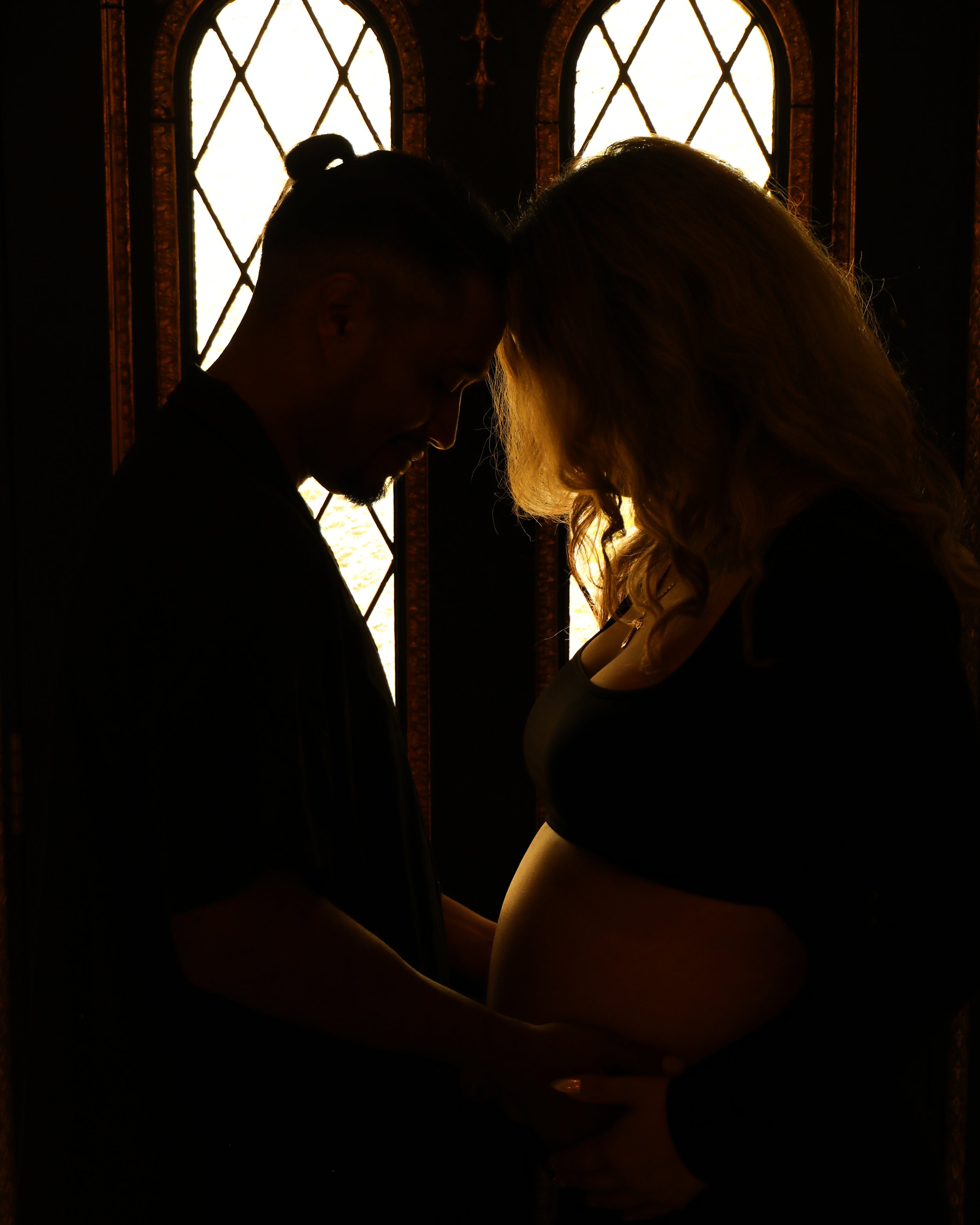 Silhouettes of two people with foreheads touching, backlit by stained glass windows, creating a warm glow around them.