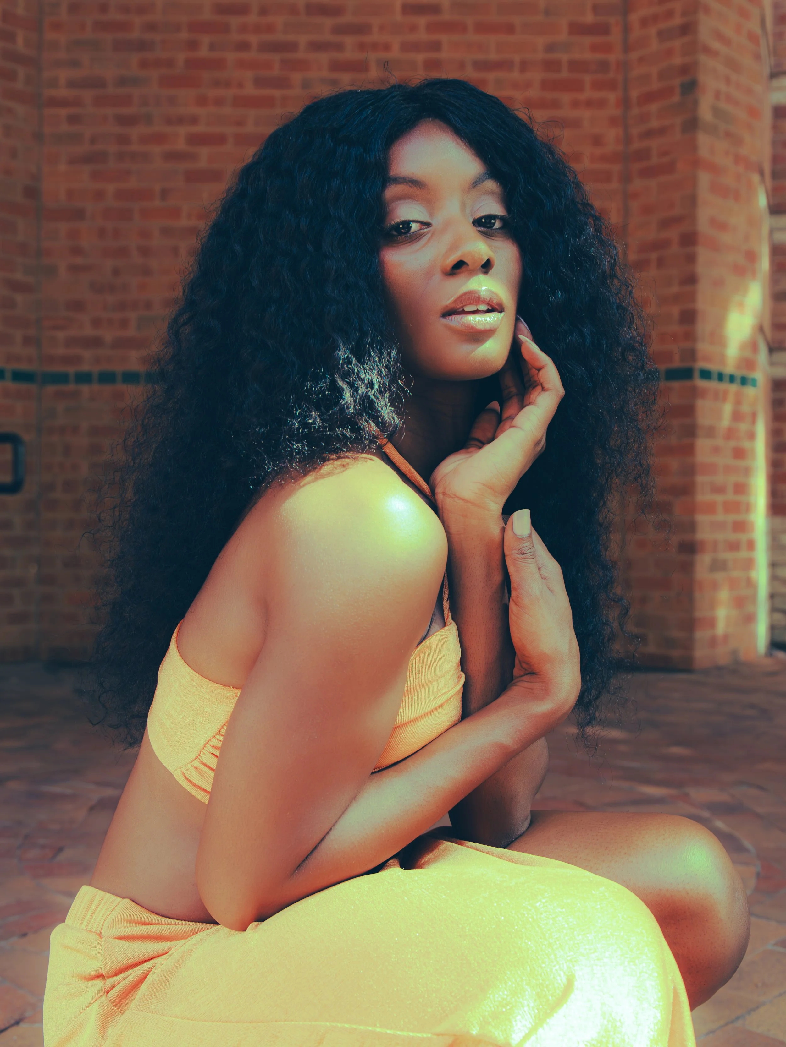 Local Dallas, Texas, DFW Non-traditional Fashion Editorial Creative Portraits Cover Art Photographer Photoshoot. A woman with curly black hair in a yellow outfit sitting against a brick wall.