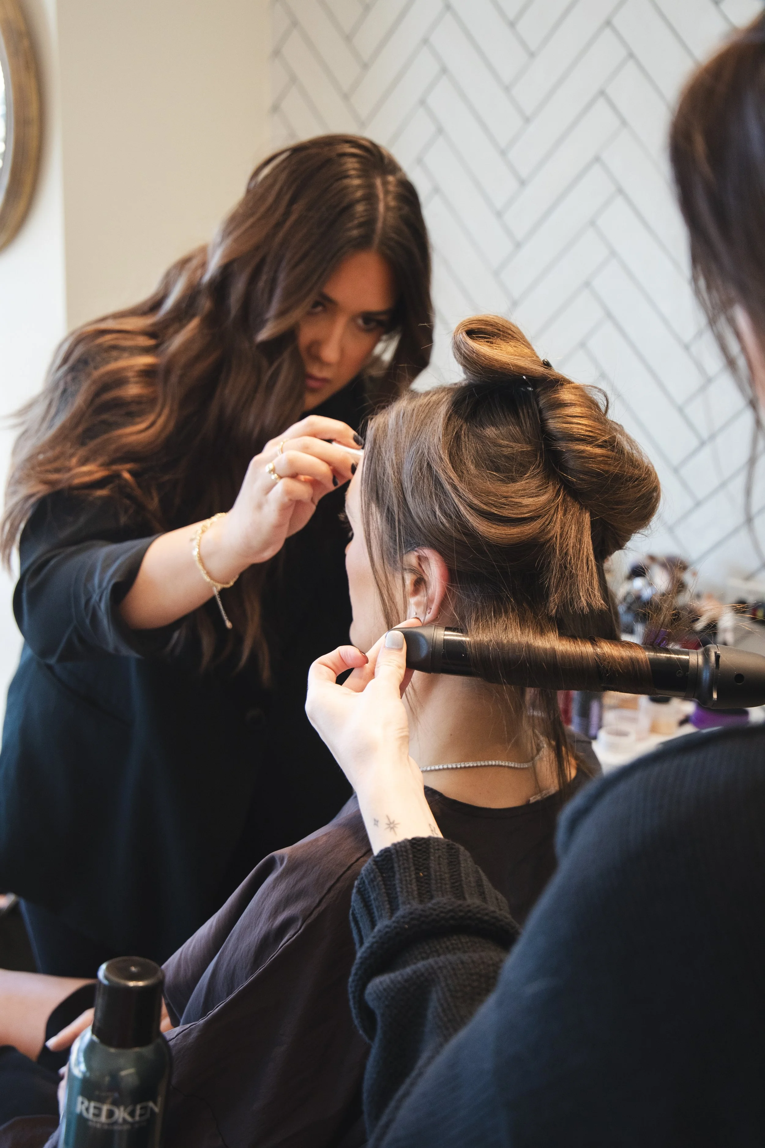 A makeup artist applying makeup to a woman. The woman is seated, and the makeup artist is focused on her face. The scene appears to be in a salon or a makeup studio, with a mirror partially visible in the background.