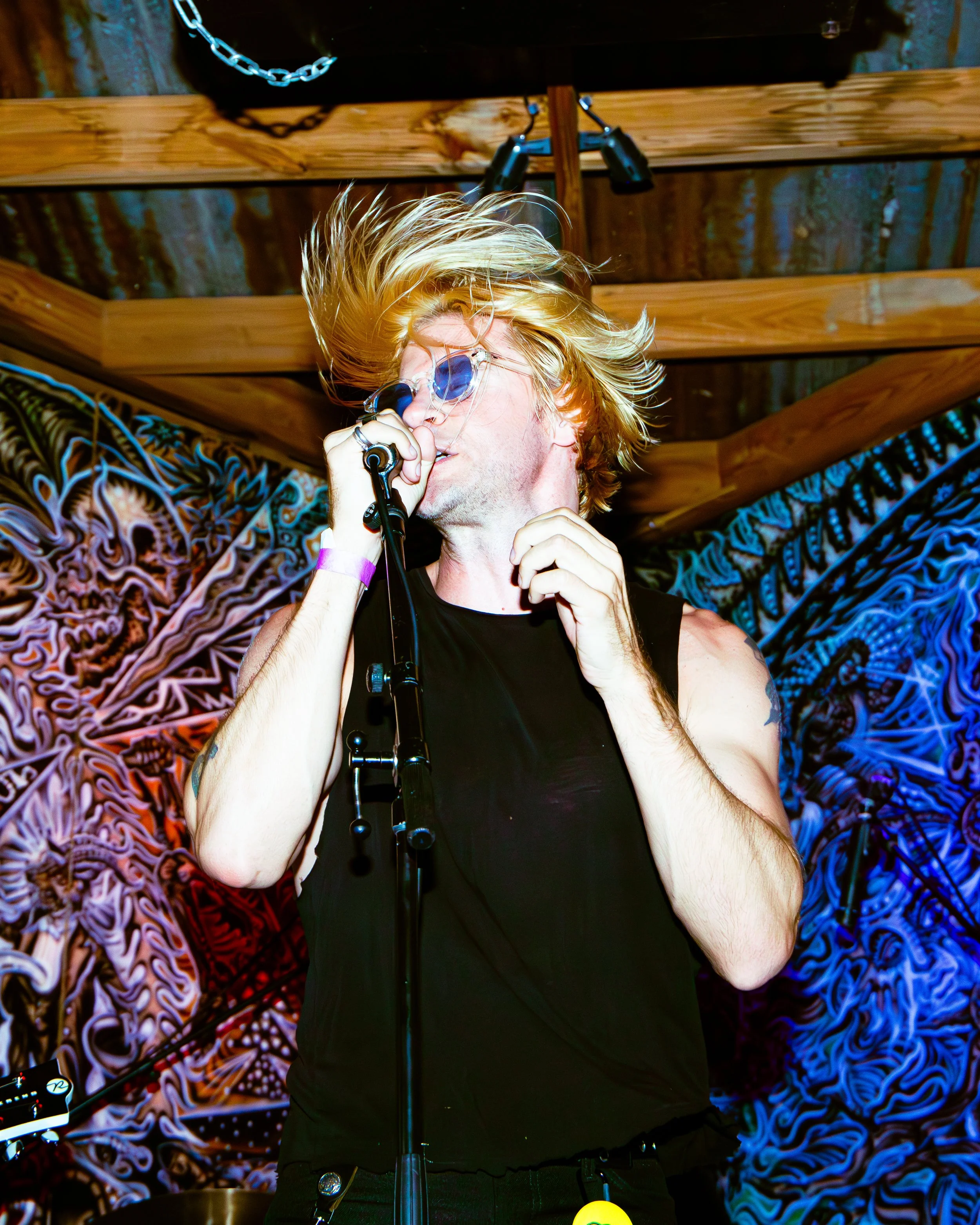 A man singing into a microphone on stage with a colorful psychedelic background and wooden beams overhead.