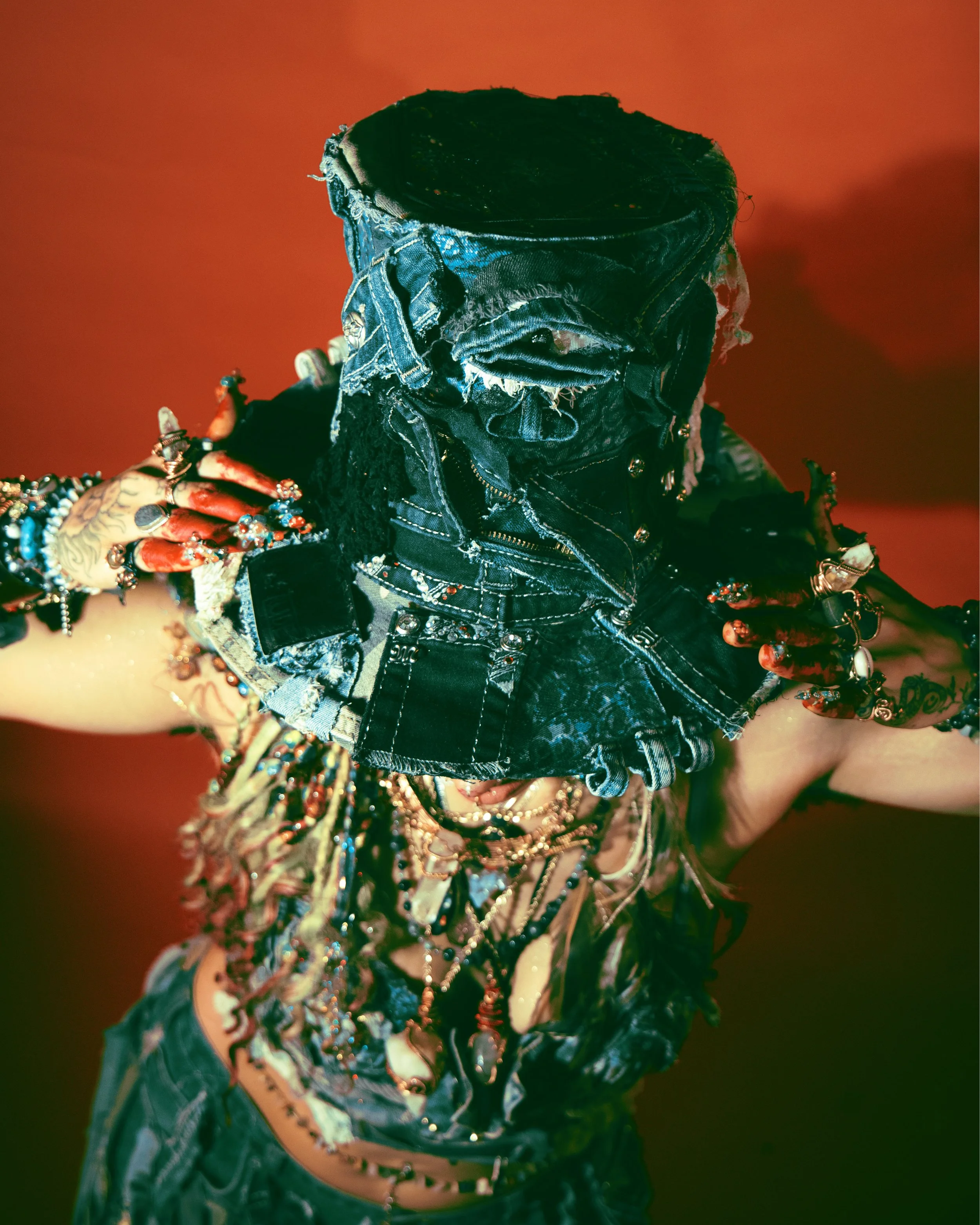Person dressed in black with colorful jewelry on an arm, holding out their hand. The background is red.