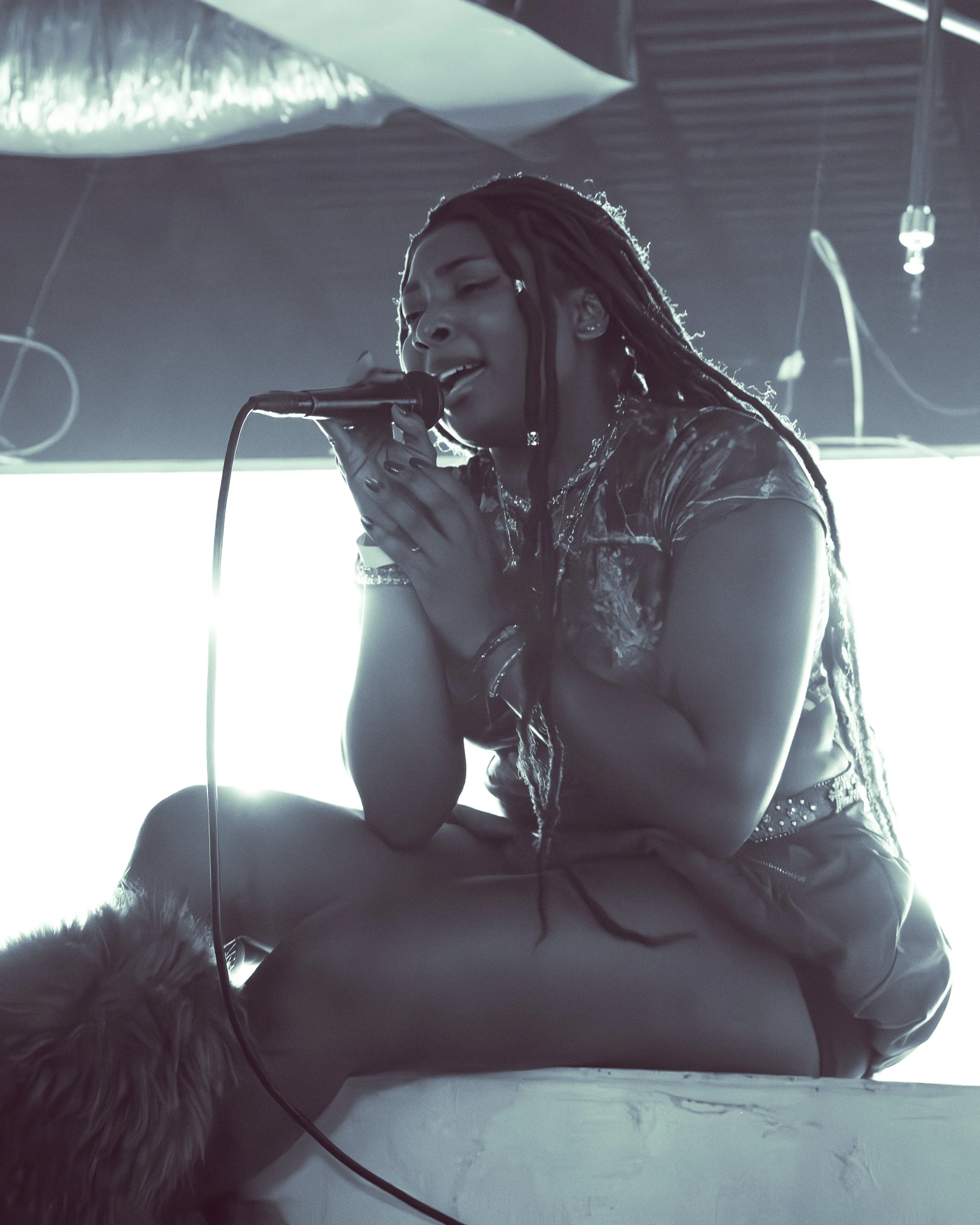 A woman with long dreadlocks singing into a microphone, with a black leather jacket, in a dimly lit setting, possibly a stage or studio.