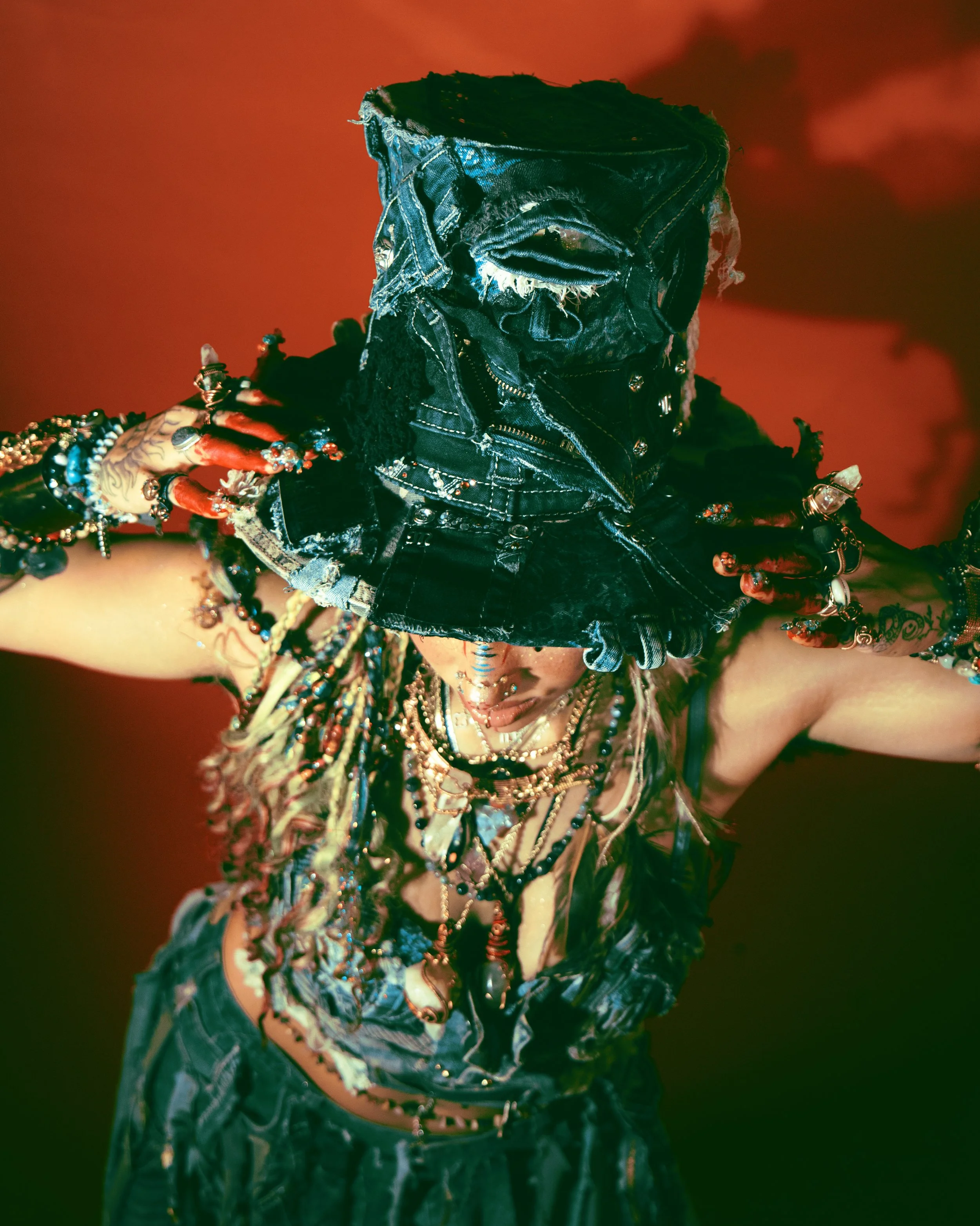 Local Dallas, Texas, DFW Non-traditional Fashion Editorial Creative Portraits Cover Art Photographer Photoshoot. @marblle._ Person wearing a denim jacket and multiple colorful bracelets, with their head covered in a denim hood, against a red backgrou