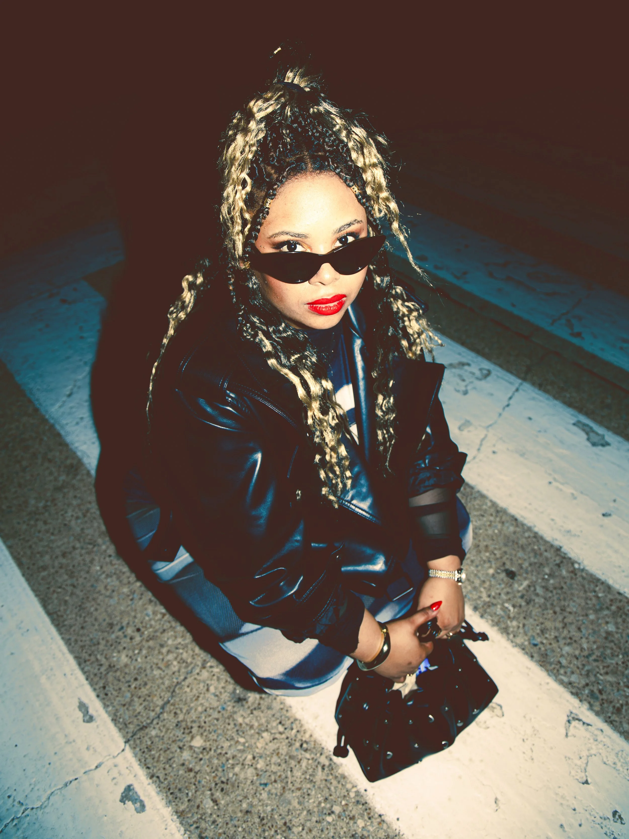 Local Dallas, Texas, DFW Non-traditional Fashion Editorial Creative Portraits Cover Art Photographer Photoshoot. A woman with long, blonde dreadlocks, wearing black sunglasses, red lipstick, a black leather jacket, and striped dress, sitting on the s