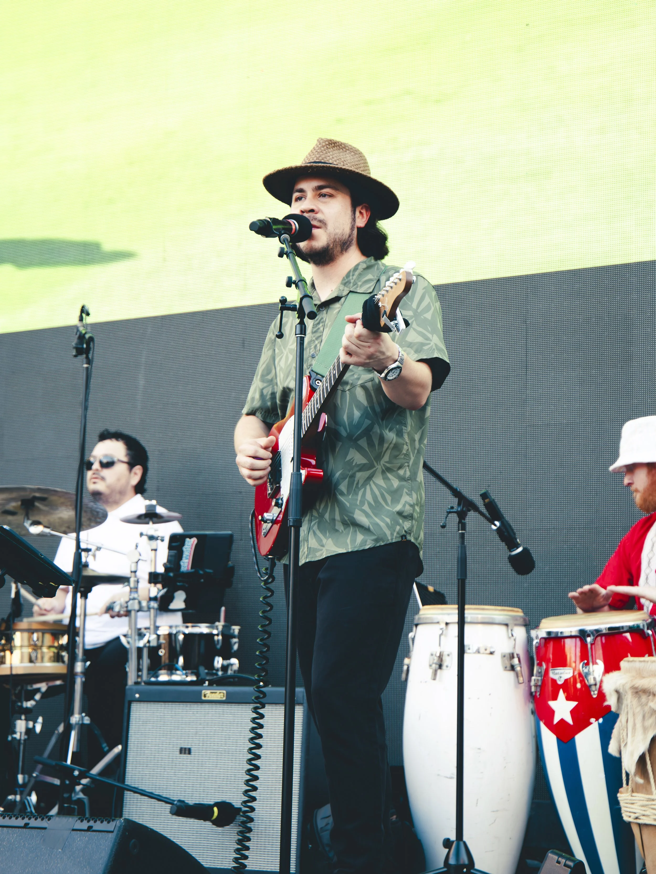 A man wearing a wide-brimmed hat and a green patterned shirt singing into a microphone and playing an electric guitar on stage. Other musicians are seen in the background with percussion instruments and conga drums.