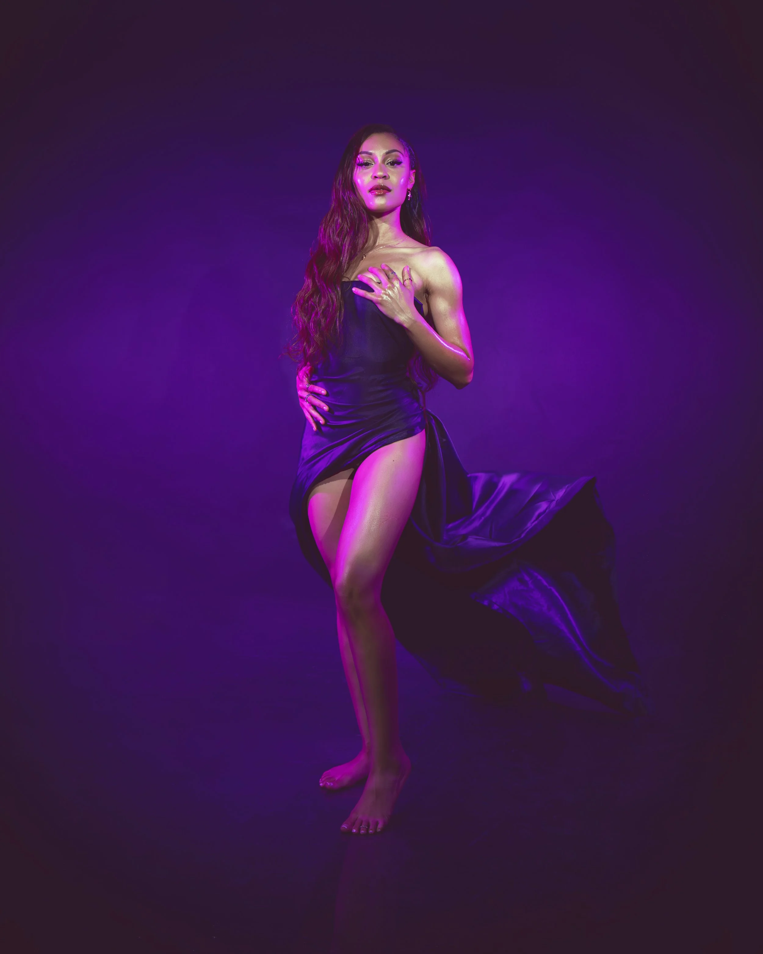 Local Dallas, Texas, DFW Non-traditional Fashion Editorial Creative Portraits Cover Art Photographer Photoshoot. Musician Rebelle Flor . A woman with long wavy hair wearing a strapless black dress standing on a dark stage illuminated with purple and 
