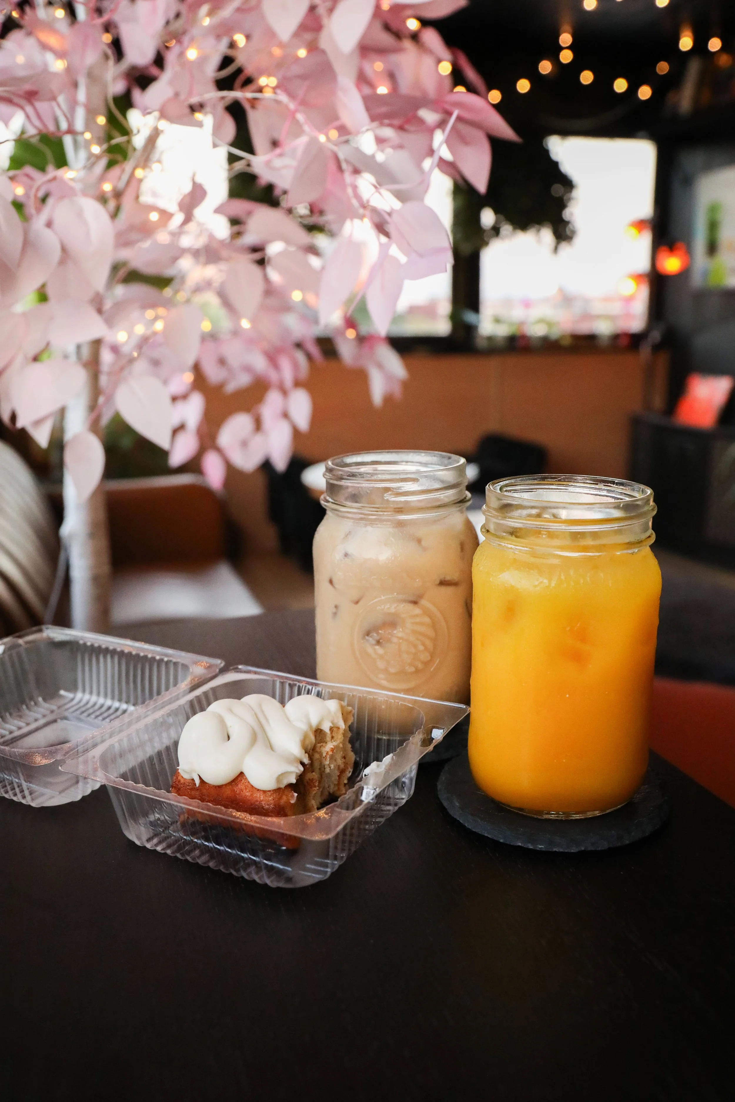 Close-up of two mason jars filled with cream and orange juice on a table, with a cupcake topped with white frosting in a clear plastic container and a pink flowering plant with string lights in the background.
