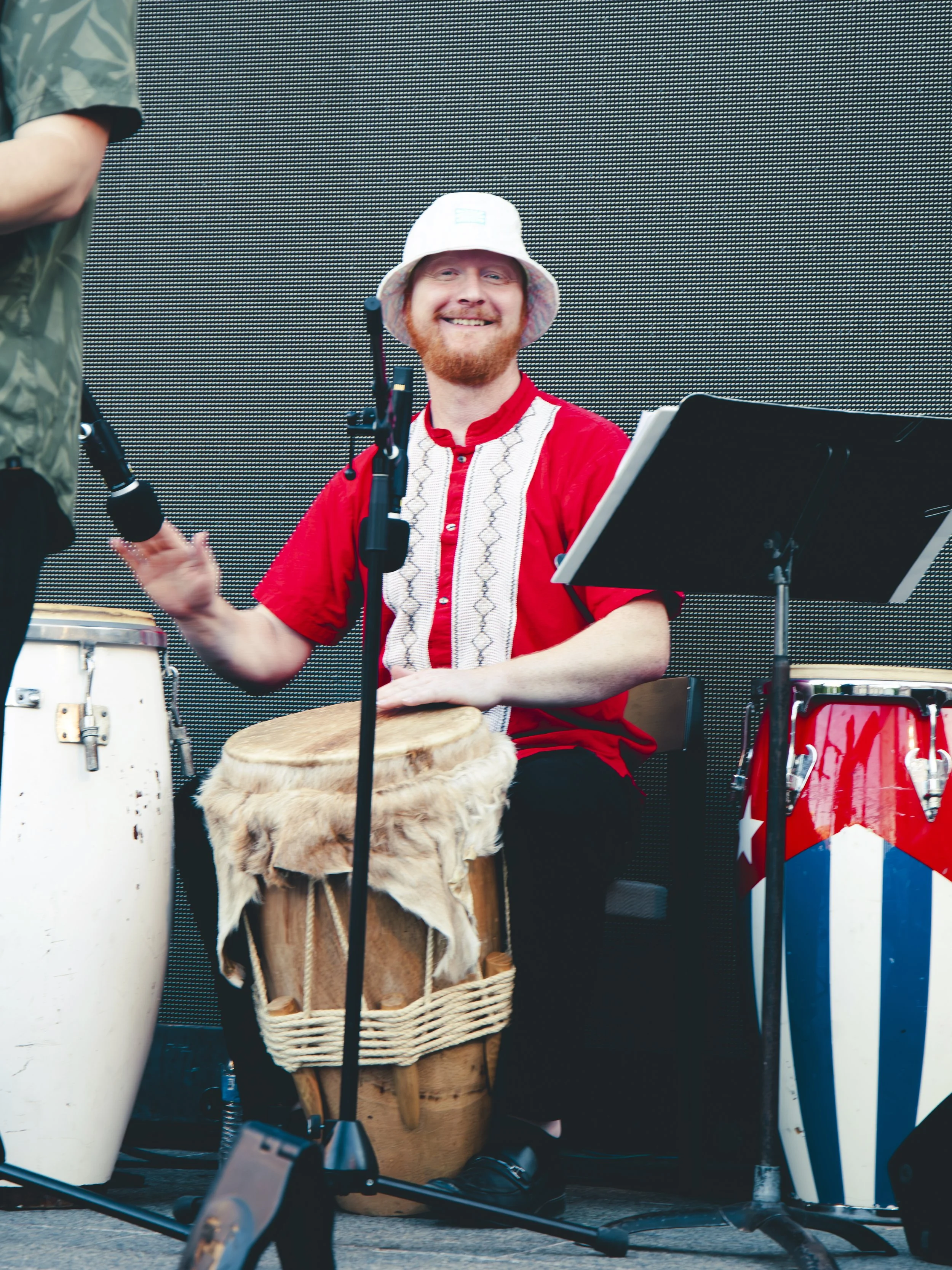 A man with a red beard smiling and playing percussion with a drum and other instruments at an outdoor event, wearing a white wide-brimmed hat and a red shirt with white embroidery.