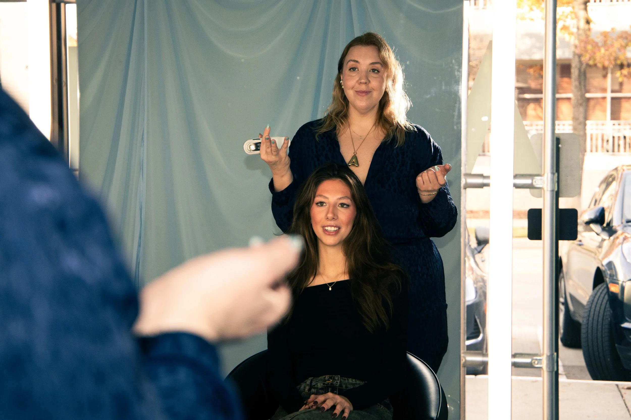 A hairstylist styling a woman's hair in a salon, with natural light coming through the windows outside.