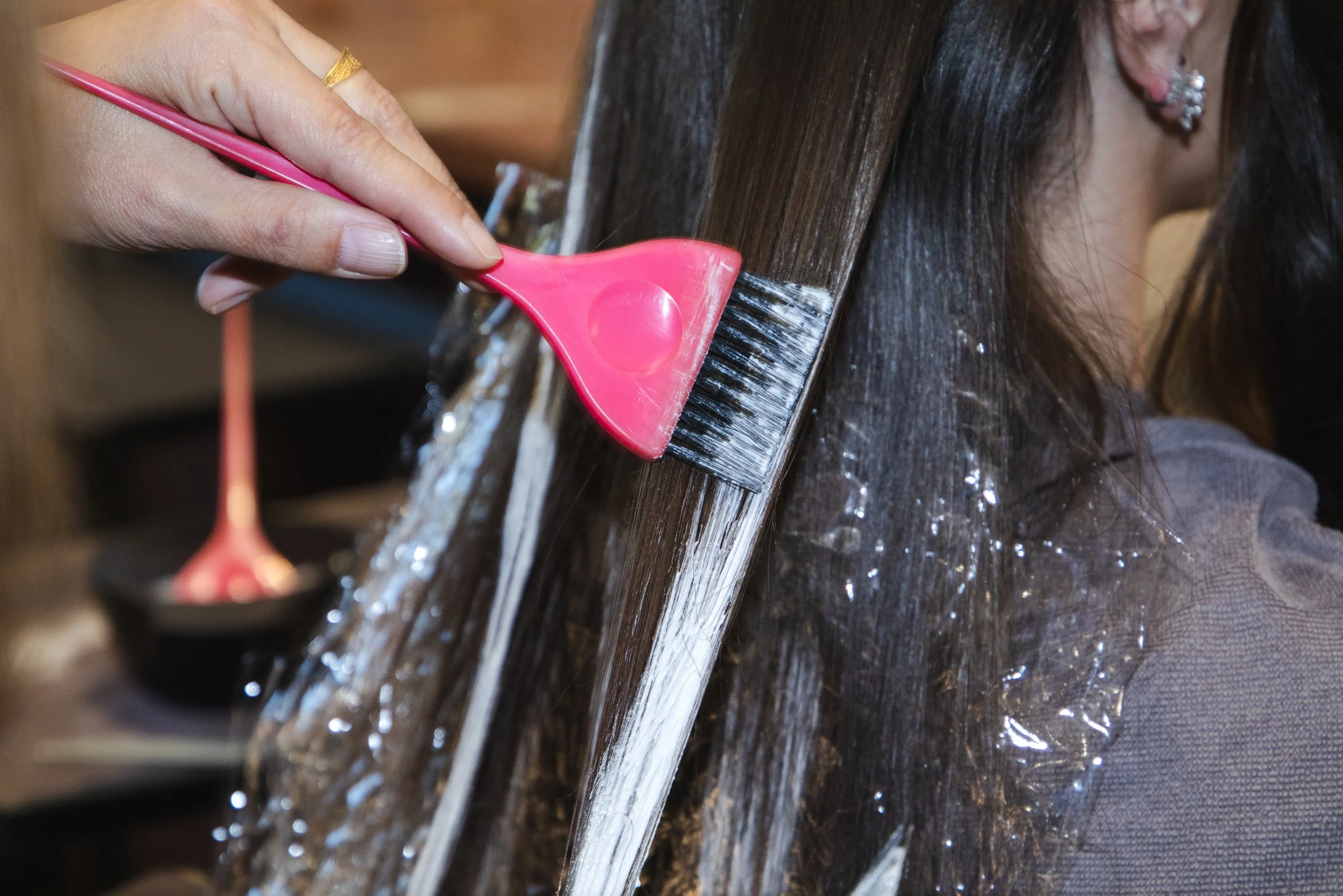 A person with earrings is using a pink silicone brush to remove white spray from dark wet hair during a salon treatment.