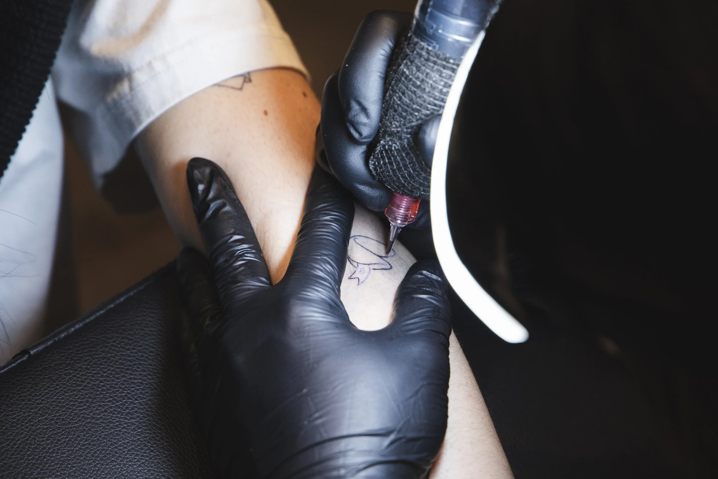 A tattoo artist wearing black gloves is creating tattoo art on a person's lower leg, which is resting on a black surface under a lamp.
