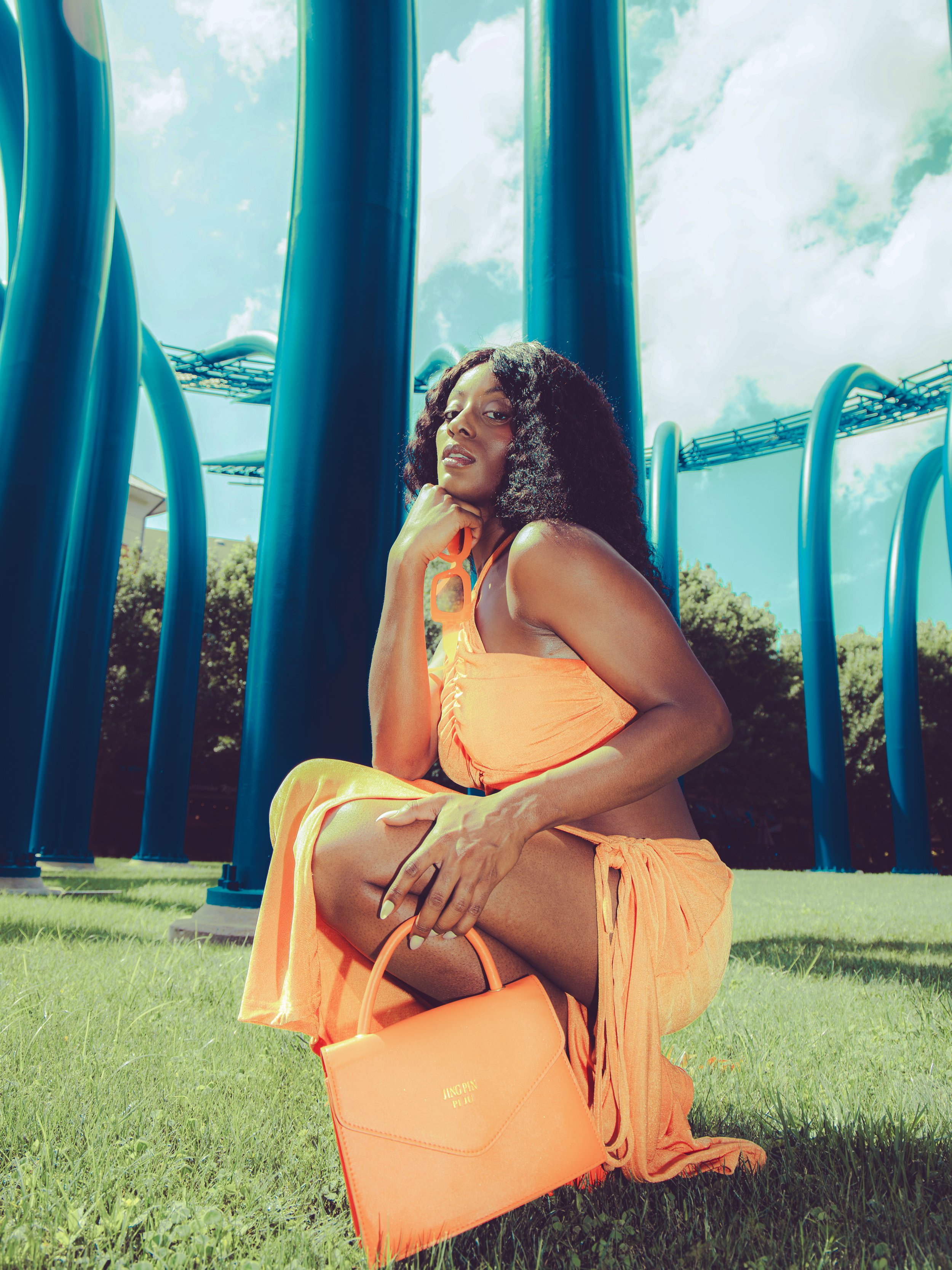 Local Dallas, Texas, DFW Non-traditional Fashion Editorial Creative Portraits Cover Art Photographer Photoshoot. A woman with curly black hair, wearing orange clothing, is sitting on green grass at a playground. She is holding an orange handbag with 