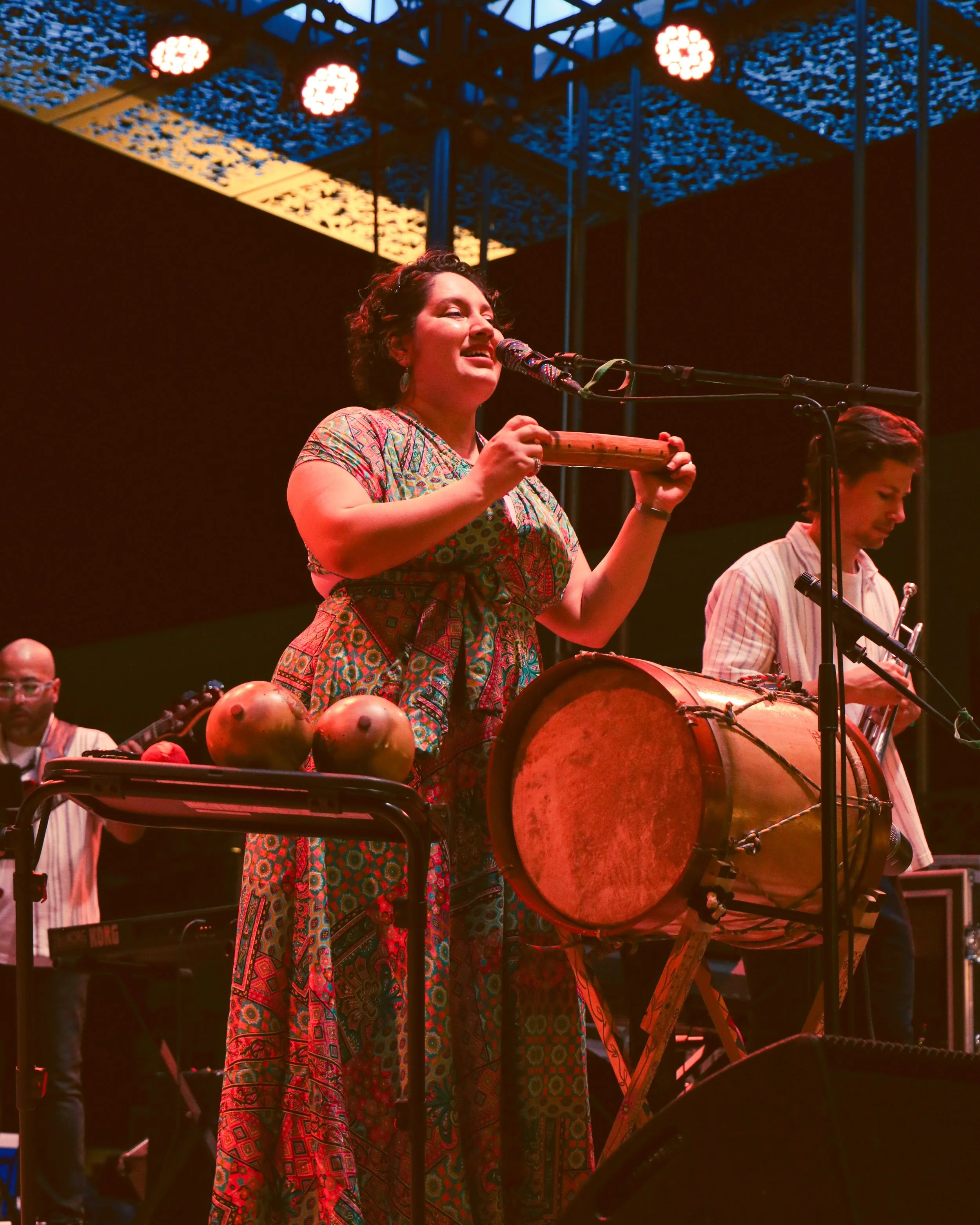 A woman with short curled hair singing and playing a traditional wooden percussion instrument at a musical performance. She is wearing a colorful, patterned dress. Behind her, a man is playing a large drum. The stage is lit with warm lighting and a b