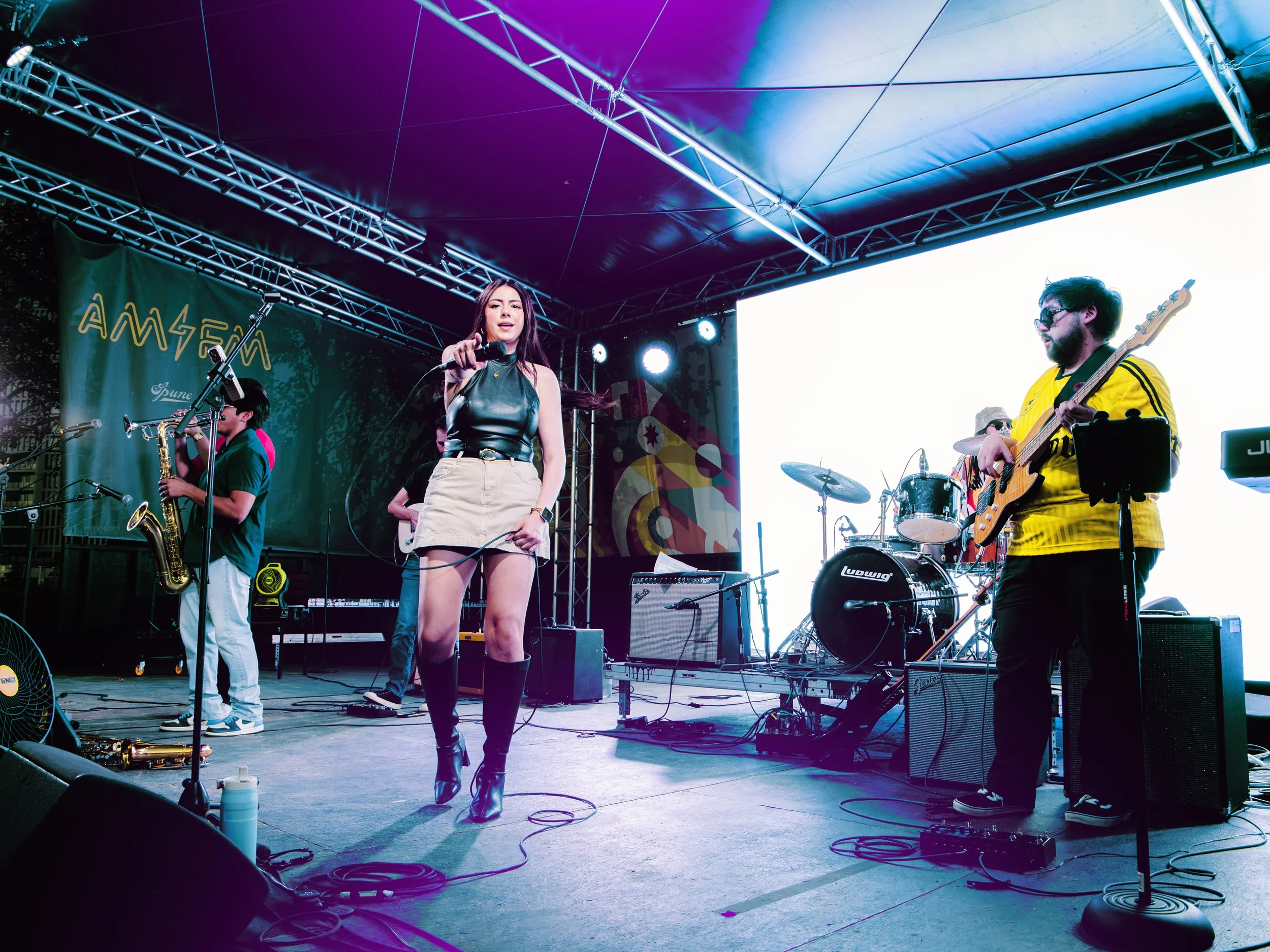 A band performing on stage outdoors with a female lead singer wearing a leather top, beige skirt, and black boots, holding a microphone. Other band members include a saxophonist and a guitarist. Stage lighting creates a colorful ambiance.