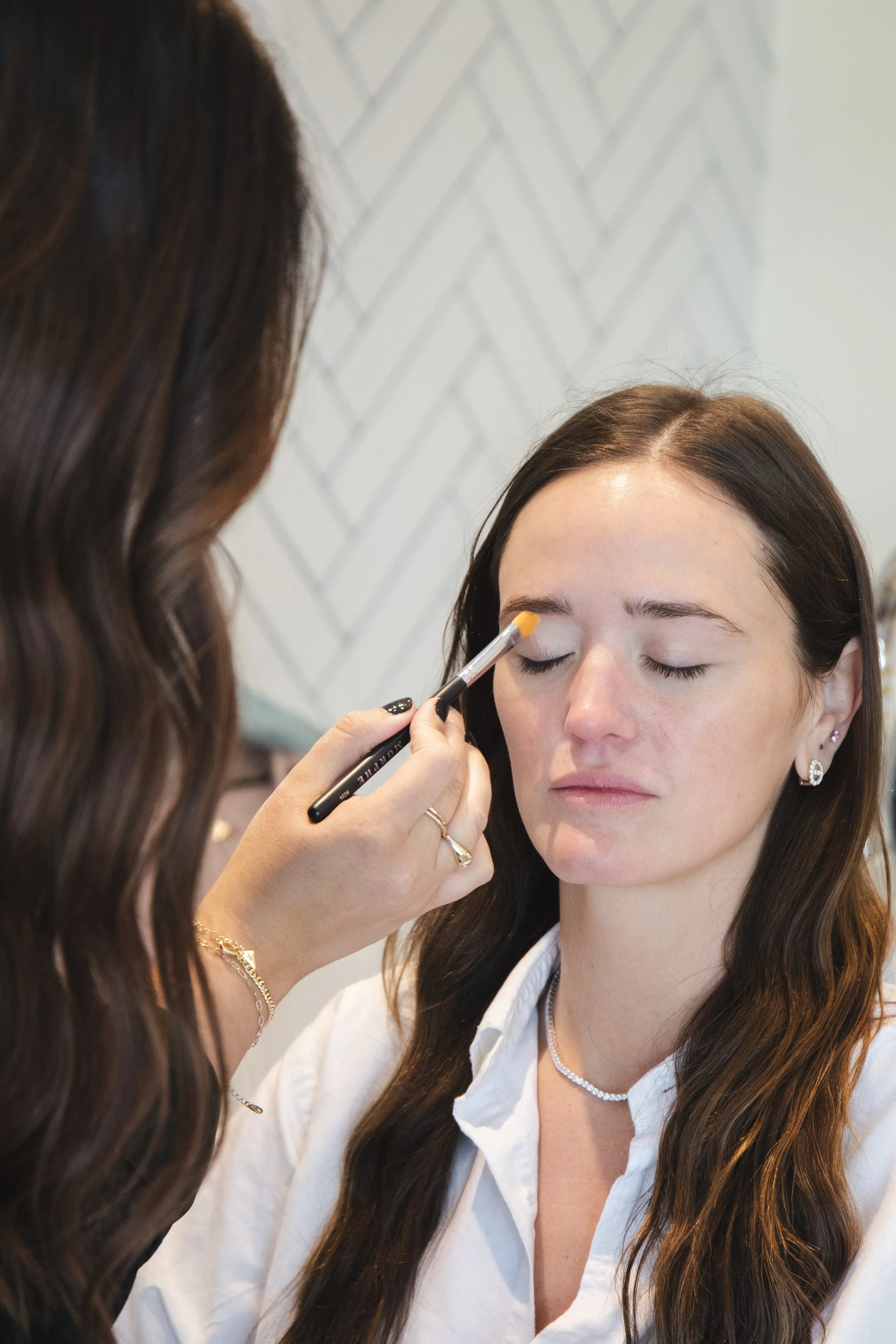 A woman applying makeup on another woman's eyebrows in a bright, modern setting.