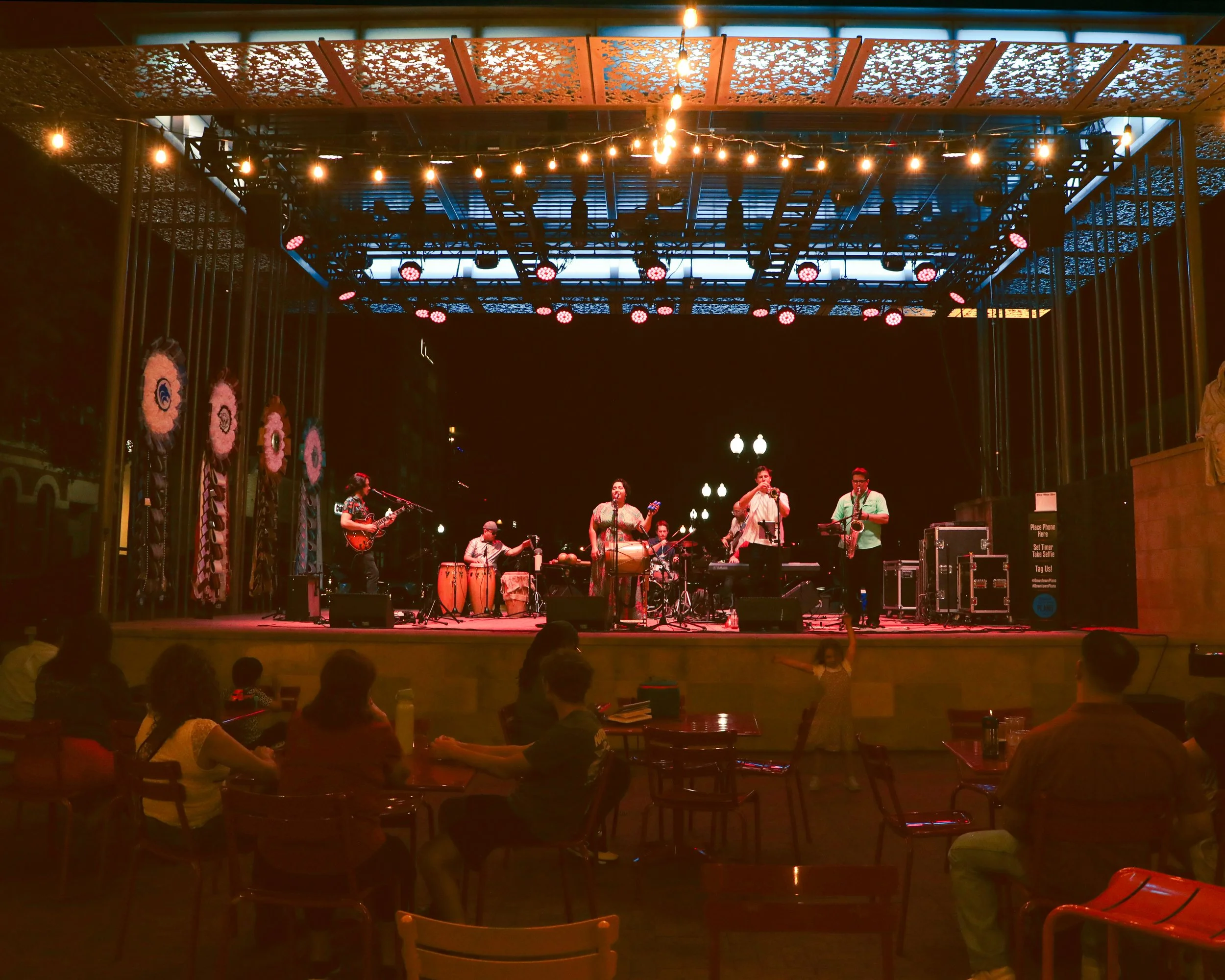 A live band performing on an outdoor stage at night. The stage has decorative elements with floral designs and string lights overhead. Audience members are seated at tables, watching the performance.