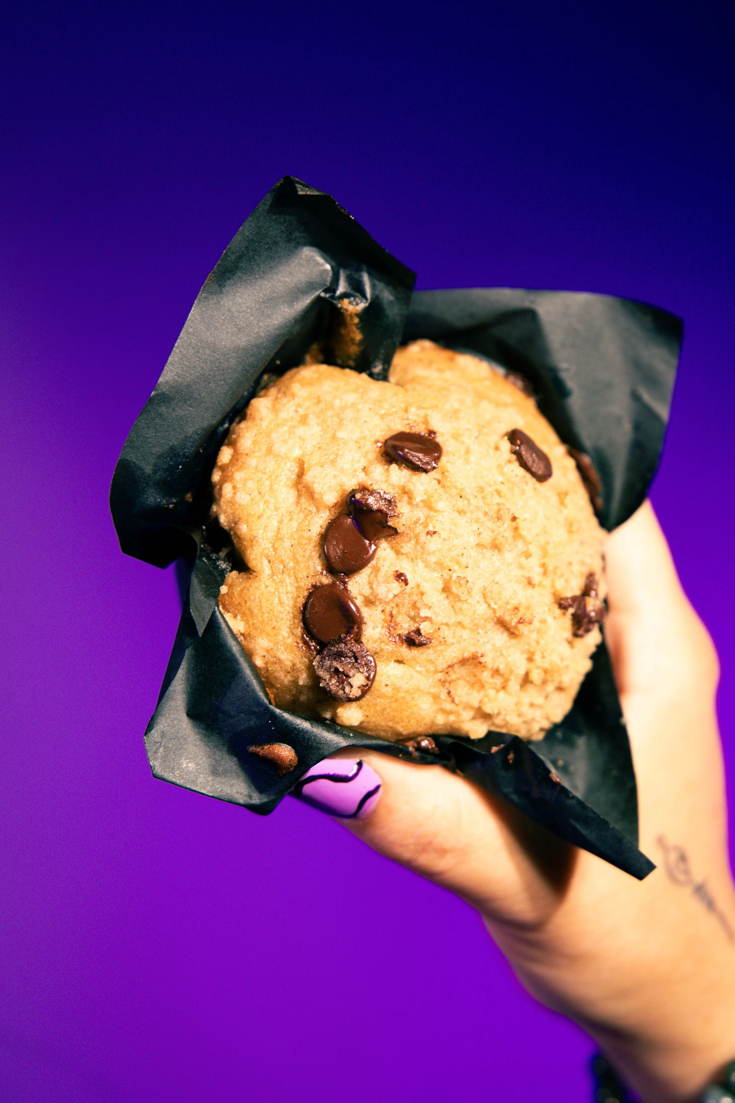 Hand holding a chocolate chip cookie in black paper wrapper against a purple background.