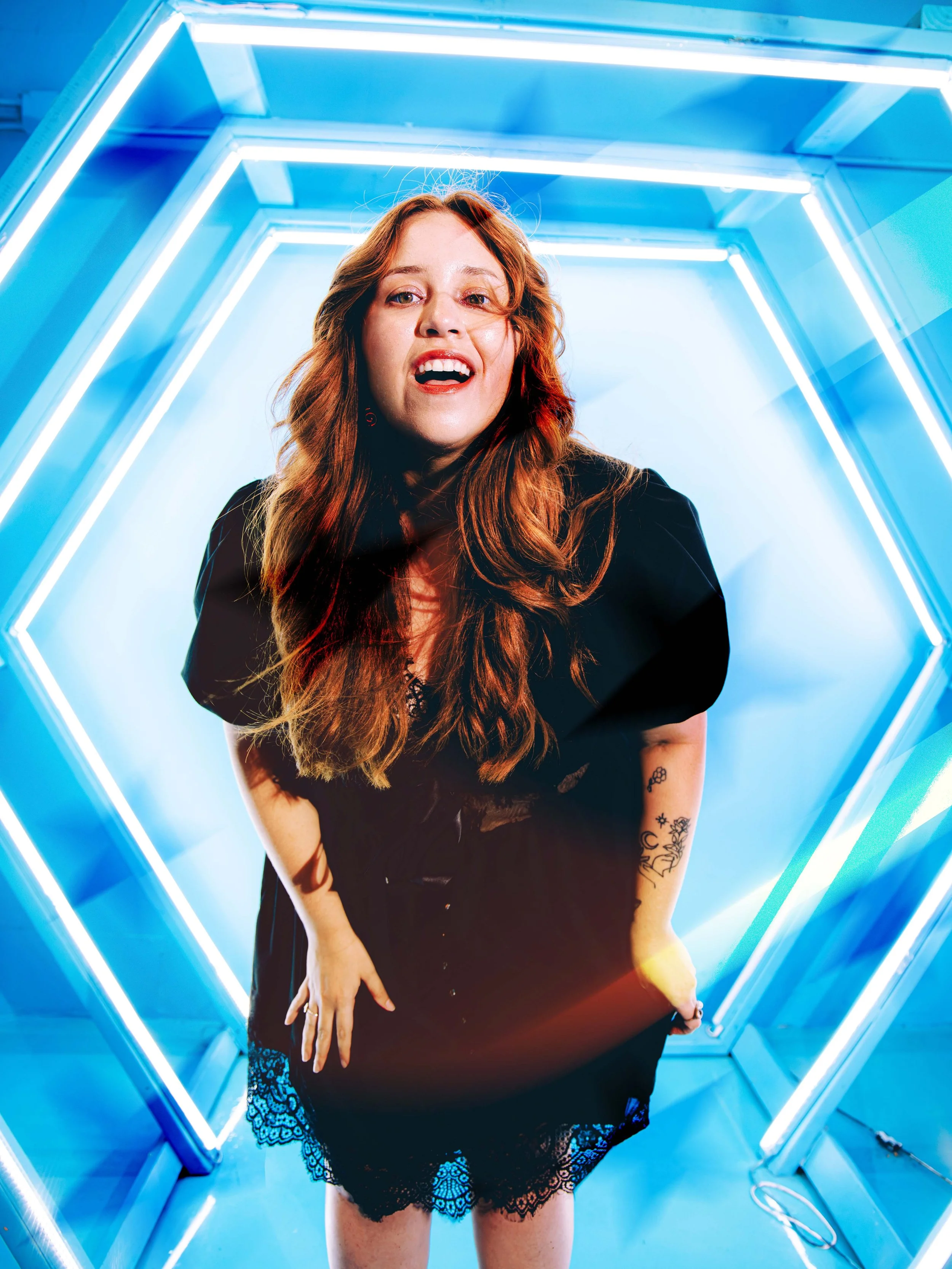 A woman with red hair and tattoos on her arms stands inside a neon-lit tunnel with blue and white lights, wearing a black dress with lace posing for a creative portrait.