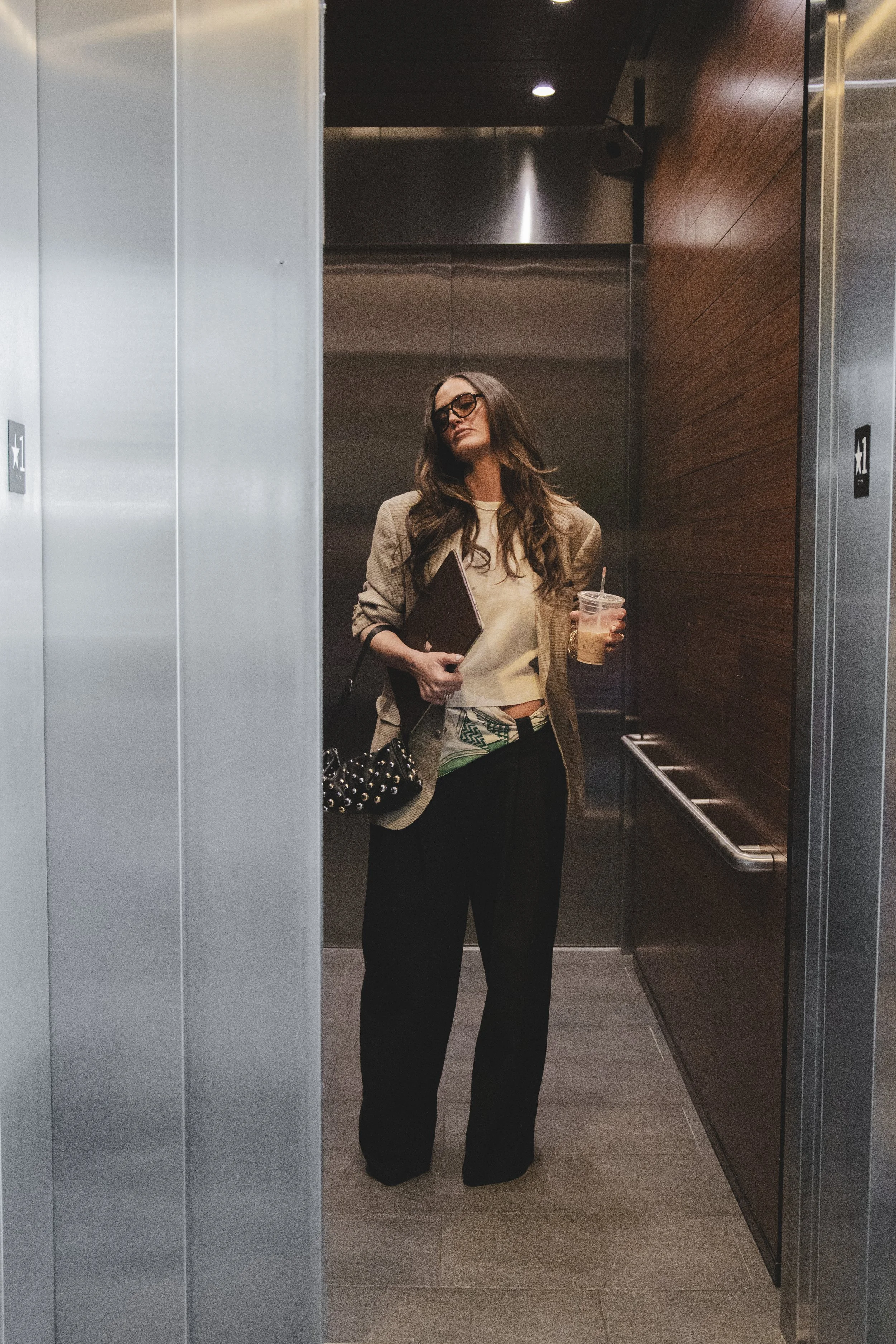 Local Dallas, Texas, DFW Non-traditional Fashion Editorial Creative Portraits Cover Art Photographer Photoshoot. Revel Workshop .A woman standing inside an elevator, holding a coffee cup and a folder, wearing glasses, a beige blazer, and black pants.