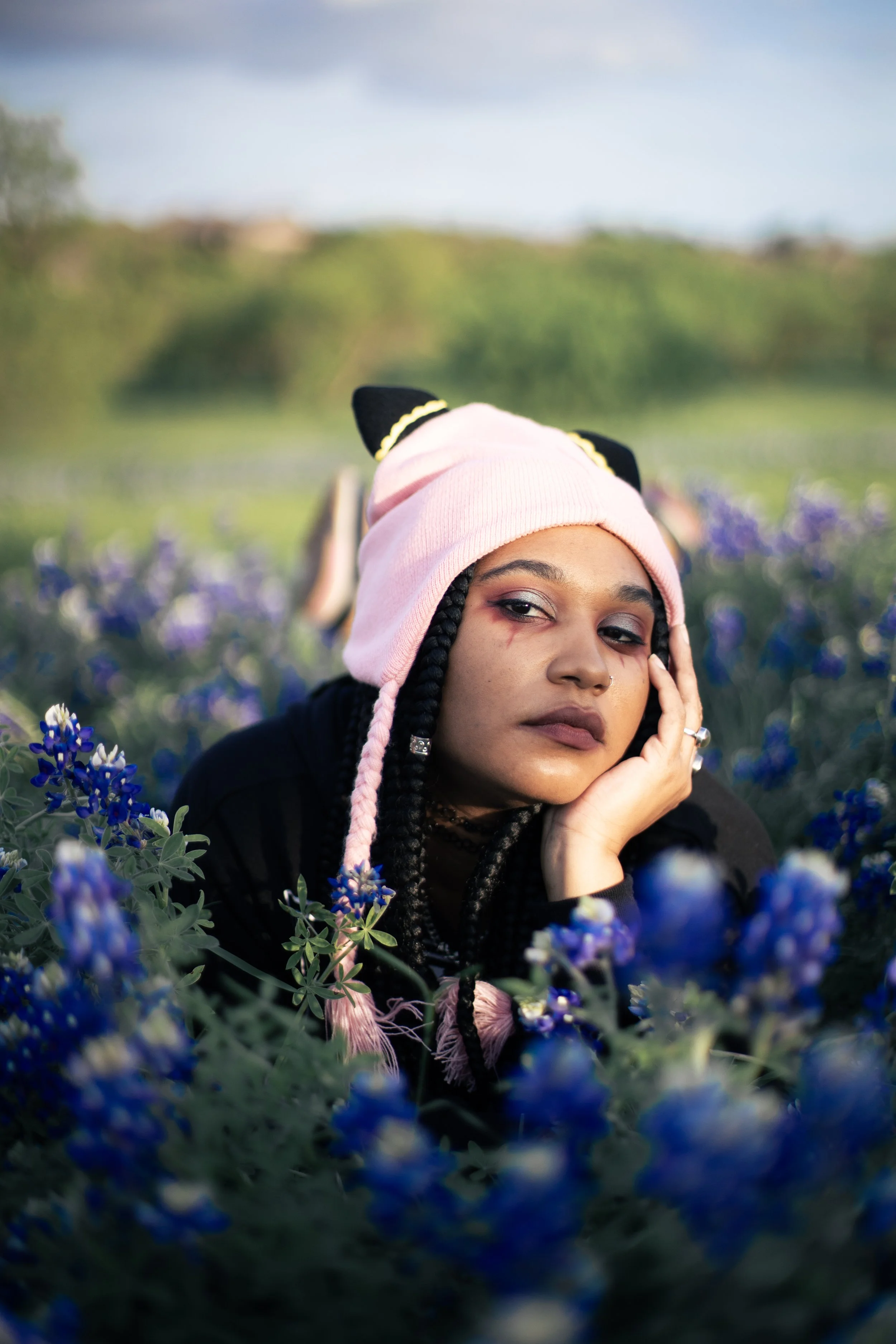 Local Dallas, Texas, DFW Non-traditional Fashion Editorial Creative Portraits Cover Art Photographer Photoshoot. musician igimeji .A young woman with braided hair, wearing a pink beanie with black and yellow accents, lying in a field of purple flower