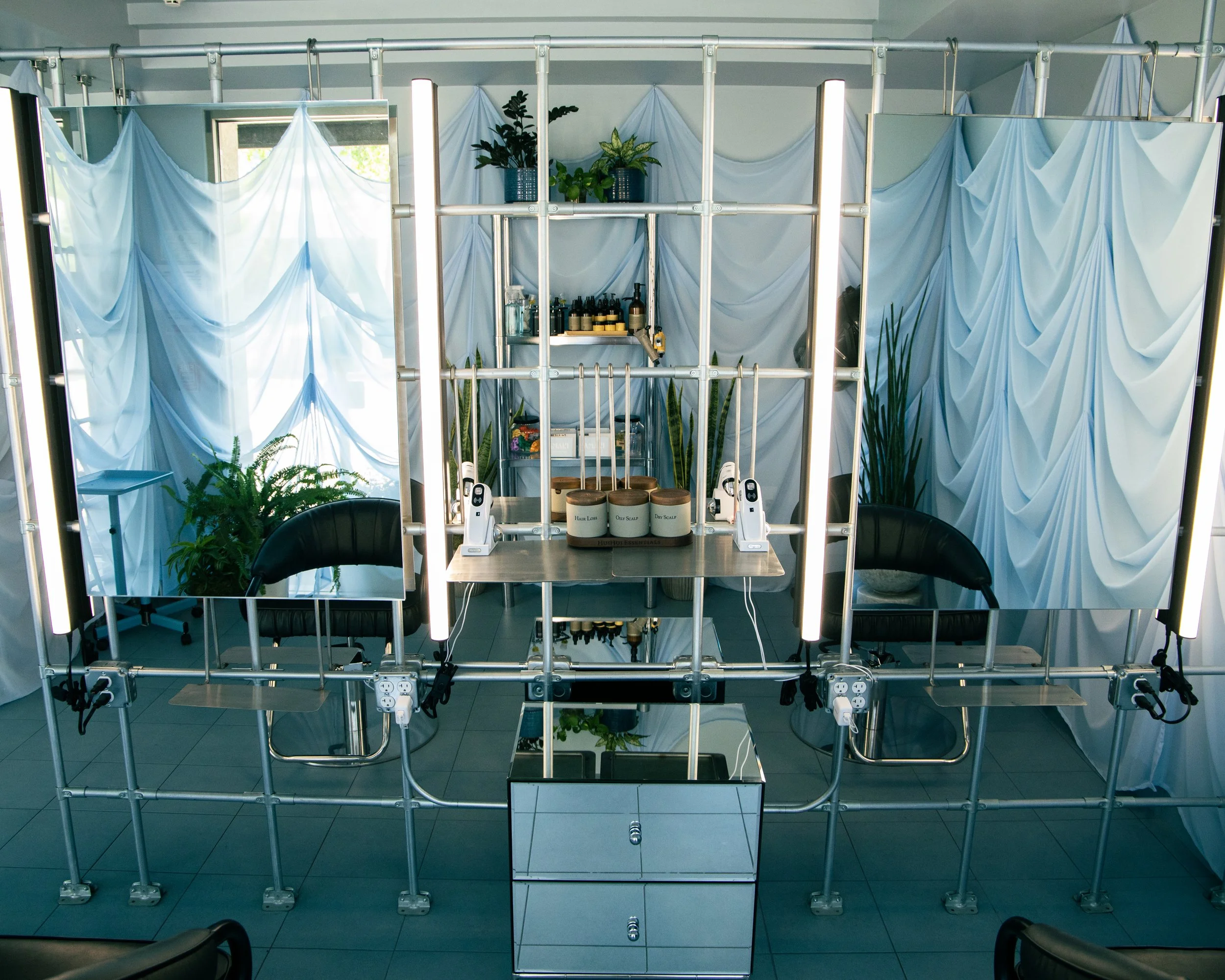A hairstyling work station with a metal frame, draped white fabric, green plants, shelves with hair products, and two styling chairs.