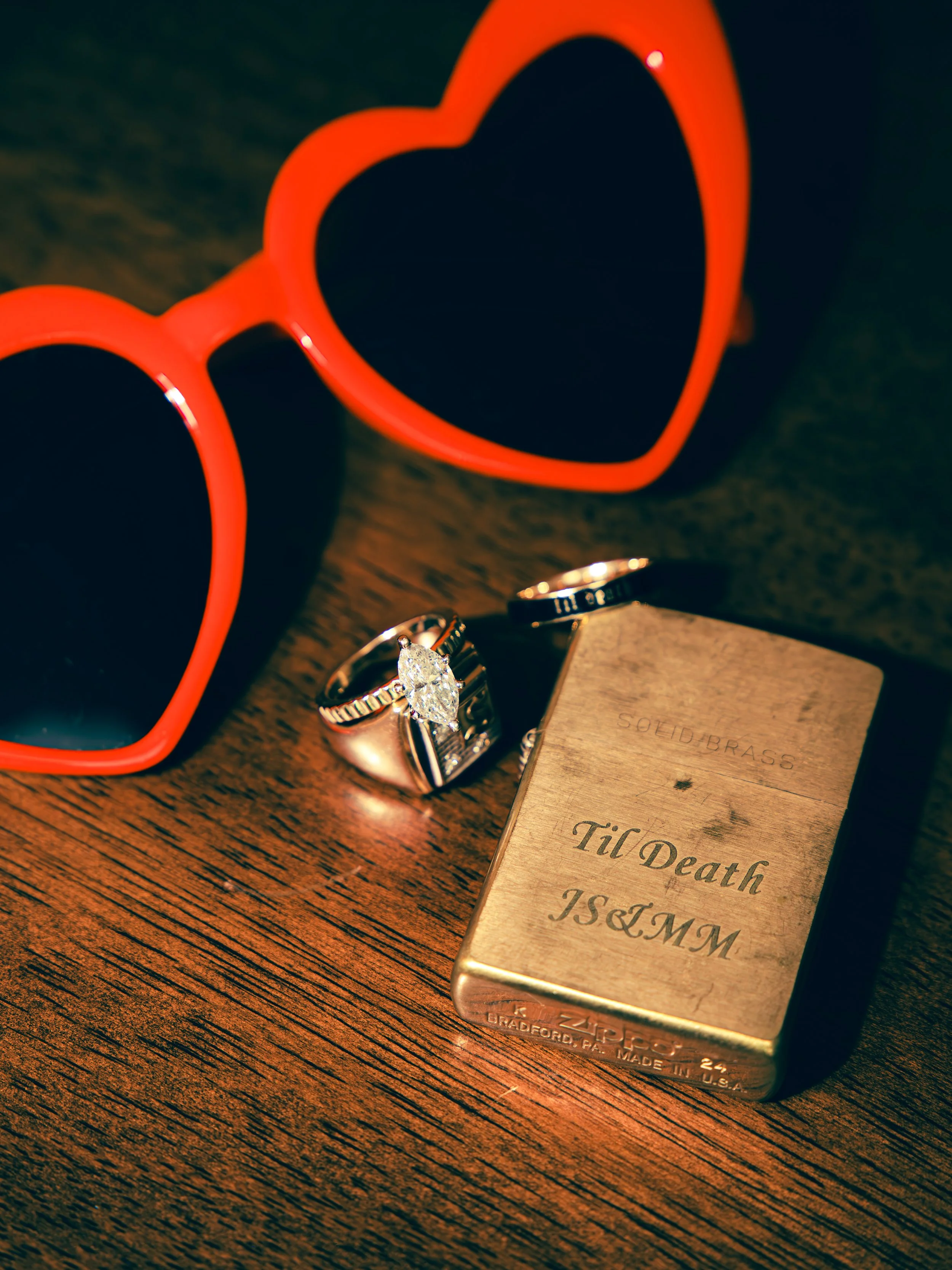 Close-up of a pair of red heart-shaped sunglasses, a diamond ring, and a gold-colored lighter on a wooden surface.