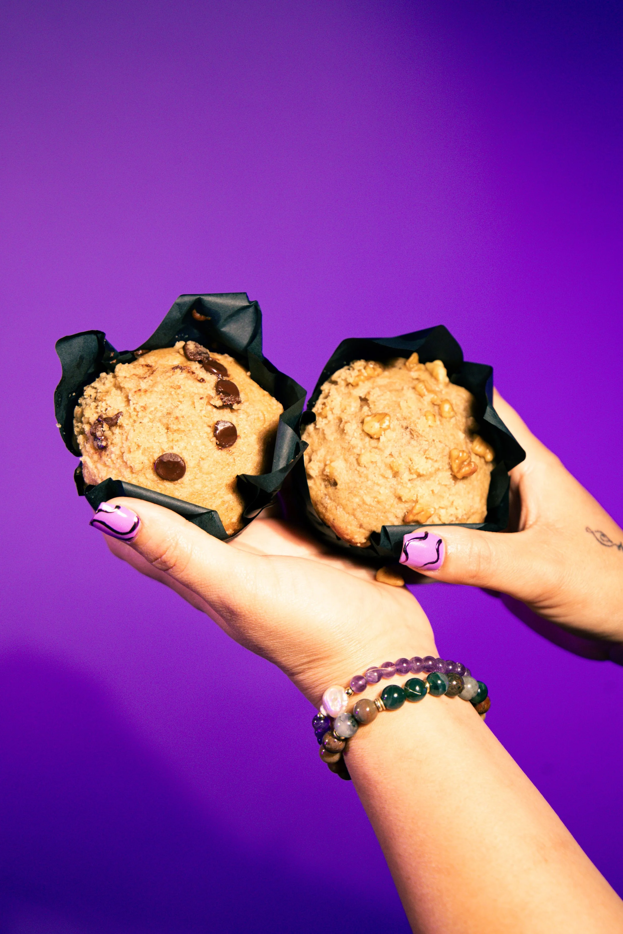 Two hands holding cookies with a purple background. The left hand has purple nail polish and wears beaded bracelets, while the right hand is visible but not wearing jewelry or nail polish.