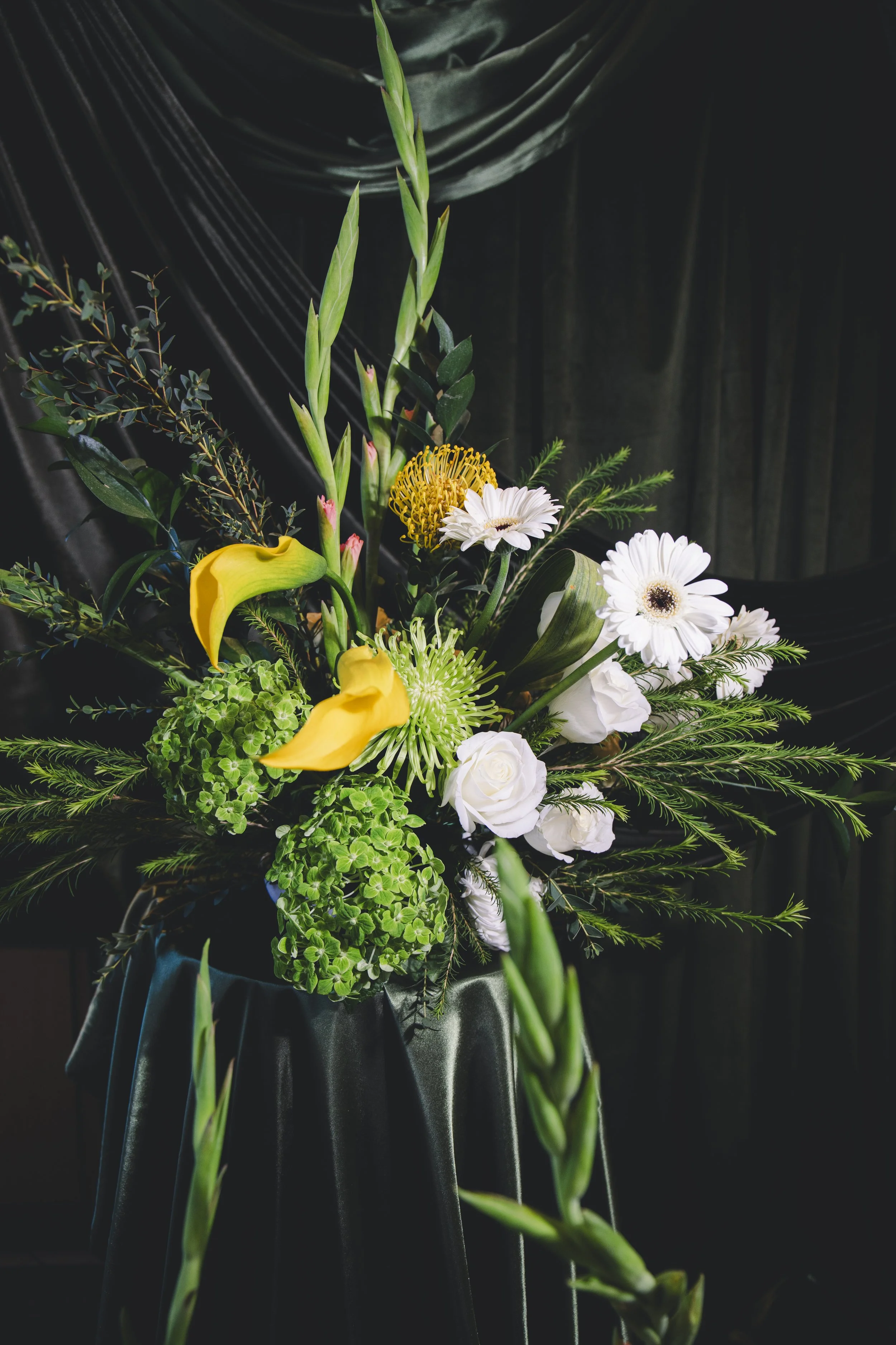 A bouquet of mixed flowers including yellow calla lilies, white daisies, green hydrangeas, and various green foliage, wrapped in black tissue paper.