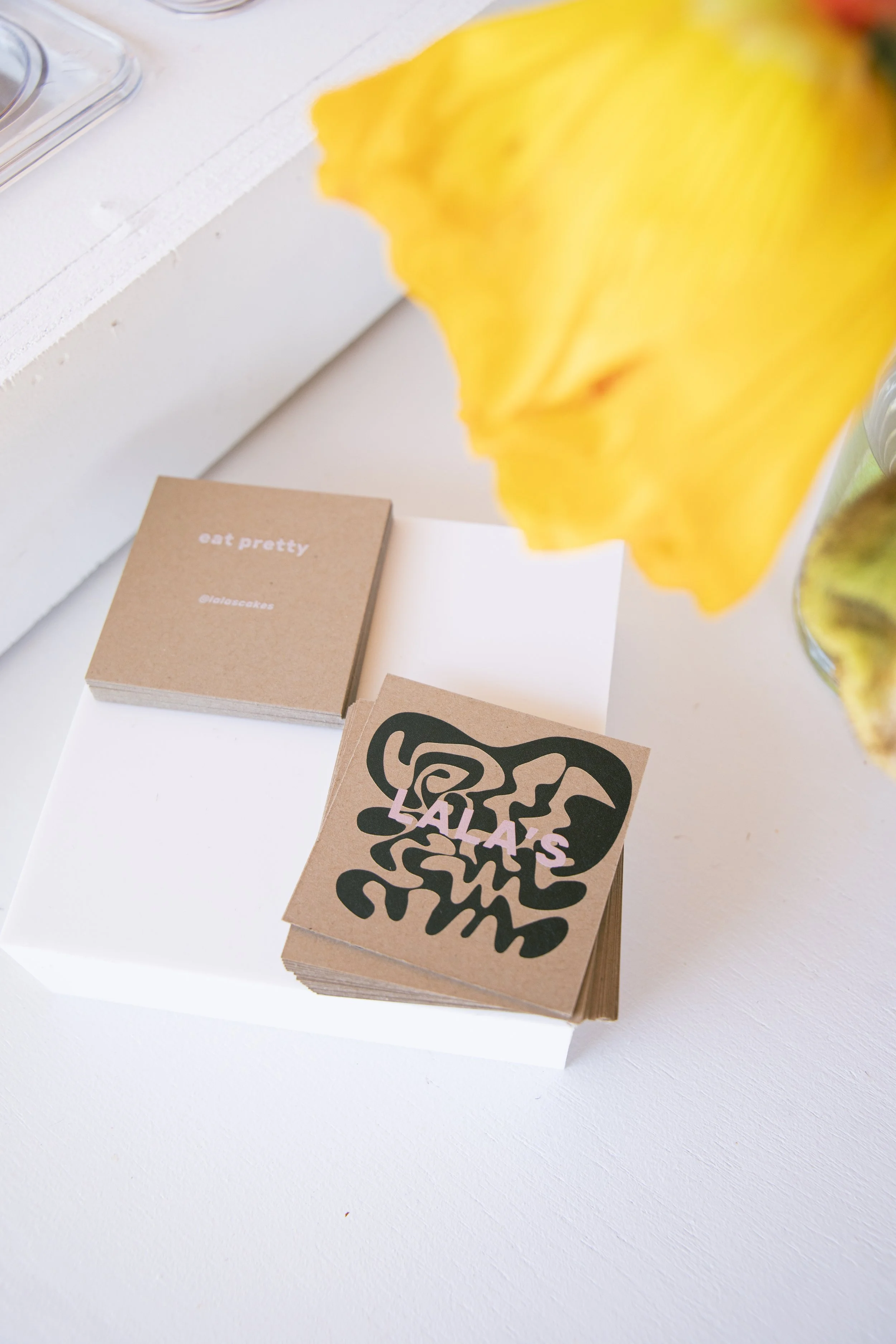 Close-up of a display with small brown paper cards, one reading "eat pretty" and another with a black graphic design, on a white surface with blurred yellow object and other items in the background.
