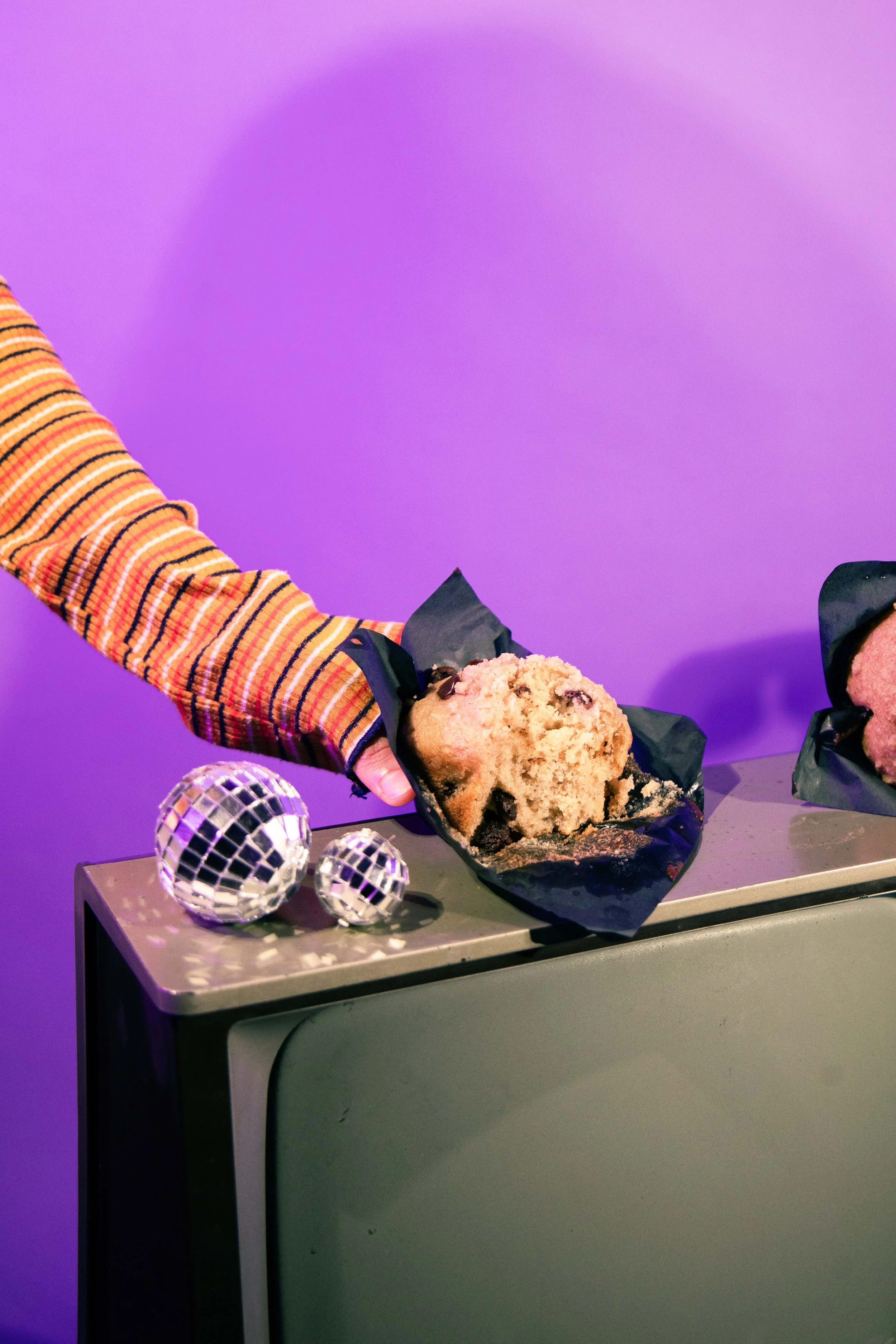 A person wearing a striped, long-sleeve shirt is reaching to pick up a large cookie with chocolate chips, wrapped in black paper, on a gray table. There are three small disco balls on the table, with a purple background.