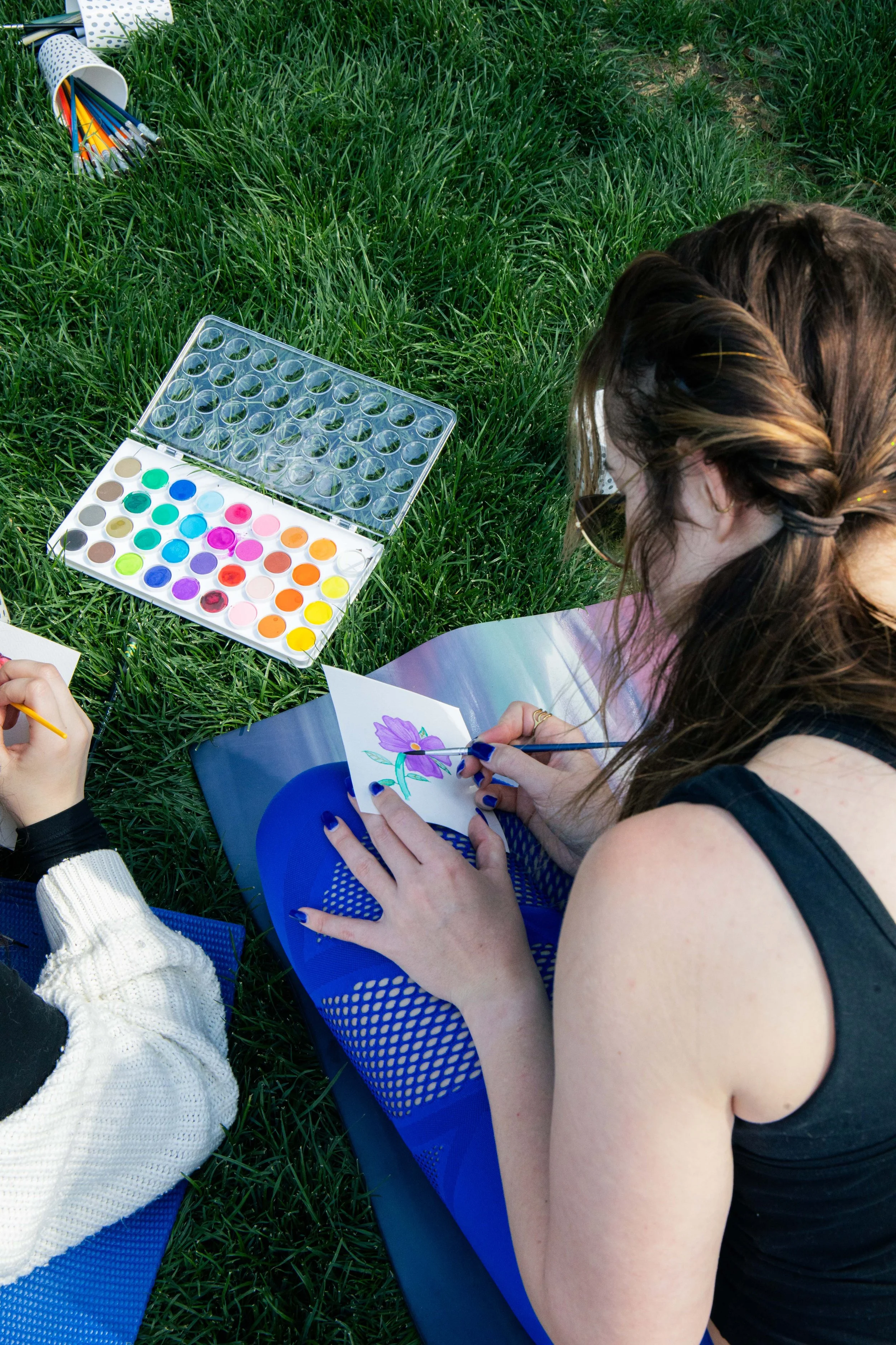 A person with brown hair and glasses sitting on the grass, painting a flower on paper with watercolor paints. Nearby are watercolor palettes, a watercolor brush, and a paper cup filled with colored pencils.