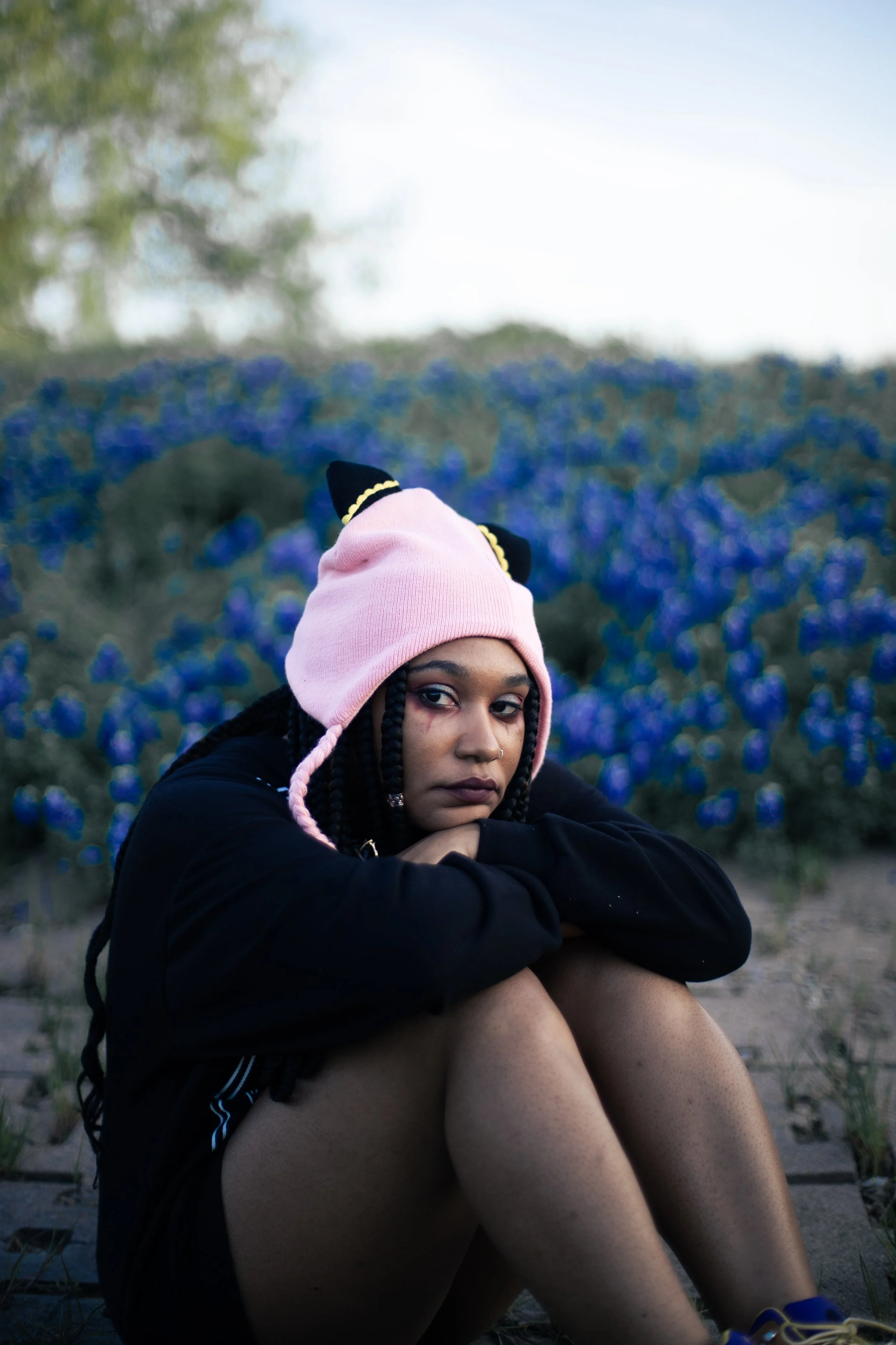 A young woman wearing a pink beanie with black ears, sitting outdoors with her arms wrapped around her knees, and looking into Local Dallas, Texas, DFW Non-traditional Fashion Editorial Creative Portraits Cover Art Photographer Photoshoot. Musician I