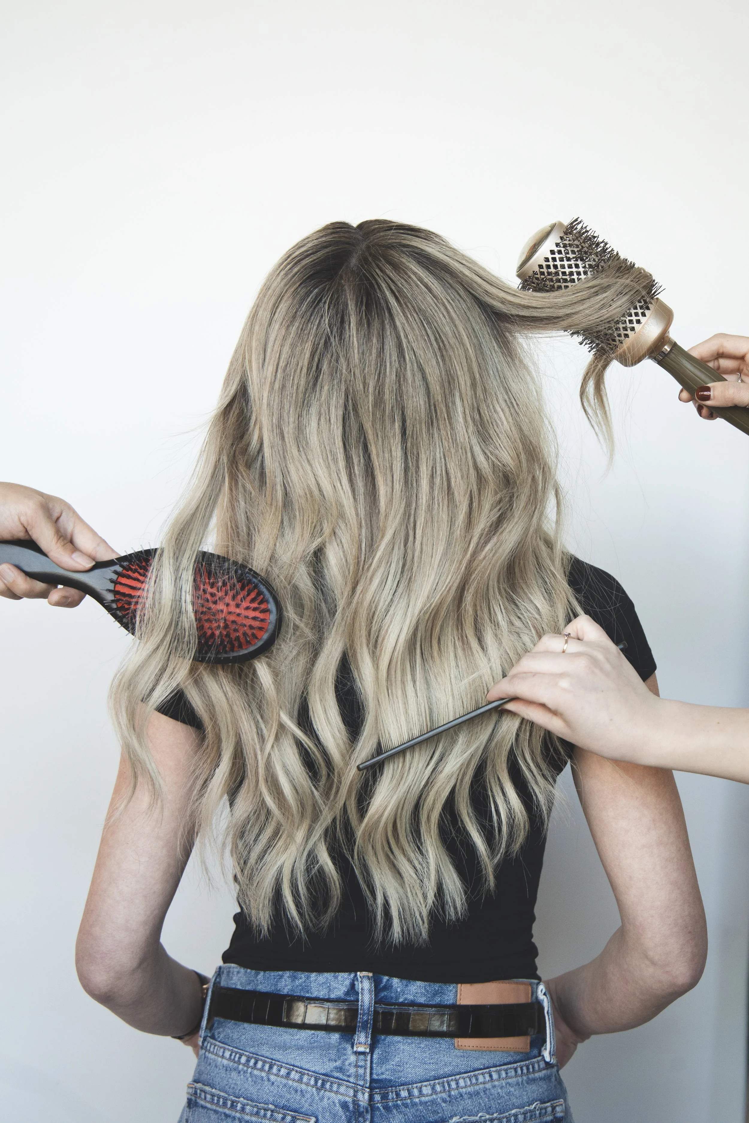 Person with long, wavy blonde hair, wearing a black shirt and high-waisted jeans, having her hair styled with a round hairbrush, a straightening iron in one hand, and a round brush in the other, against a plain white background.