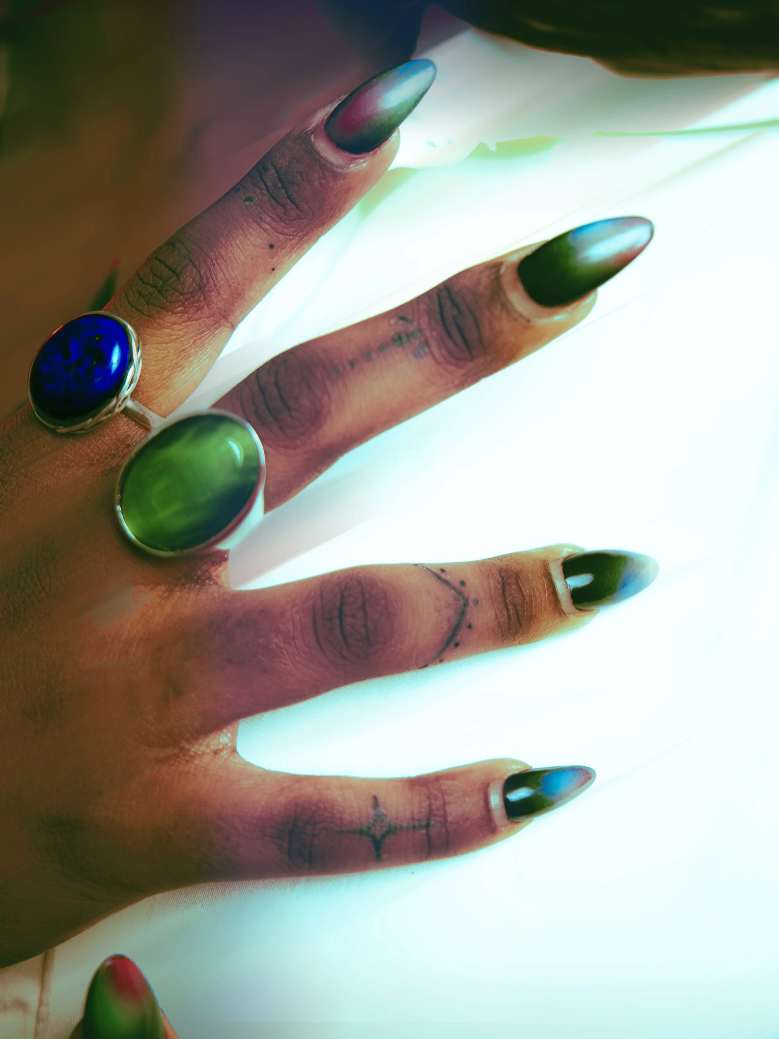 Close-up of a hand with brightly colored, long, pointed nails and two large rings with green and blue stones, resting on a glowing surface.