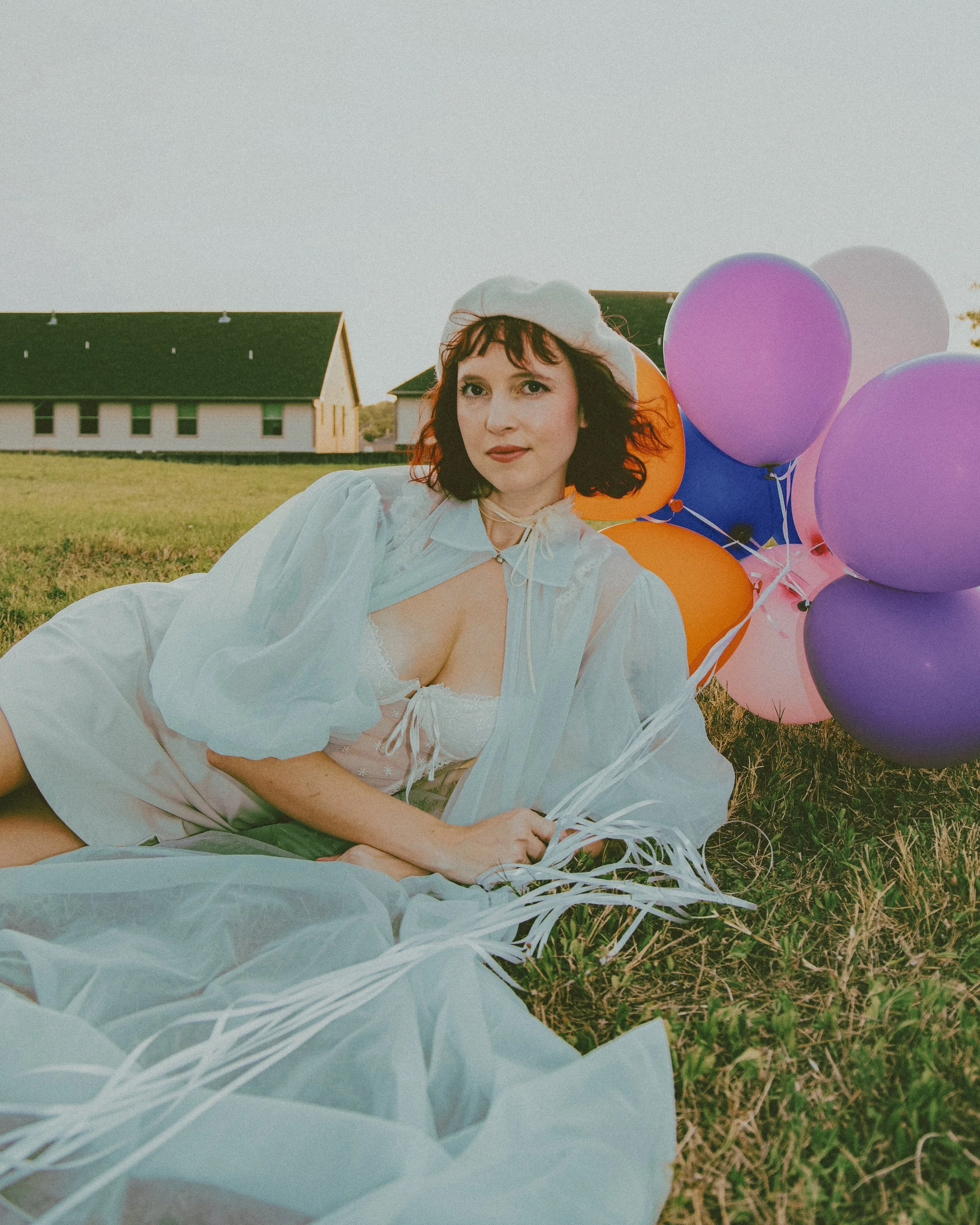 Local Dallas, Texas, DFW Non-traditional Fashion Editorial Creative Portraits Cover Art Photographer Photoshoot. A woman in a white dress and bonnet lying on grass with a bunch of colorful balloons, with houses in the background during the evening.