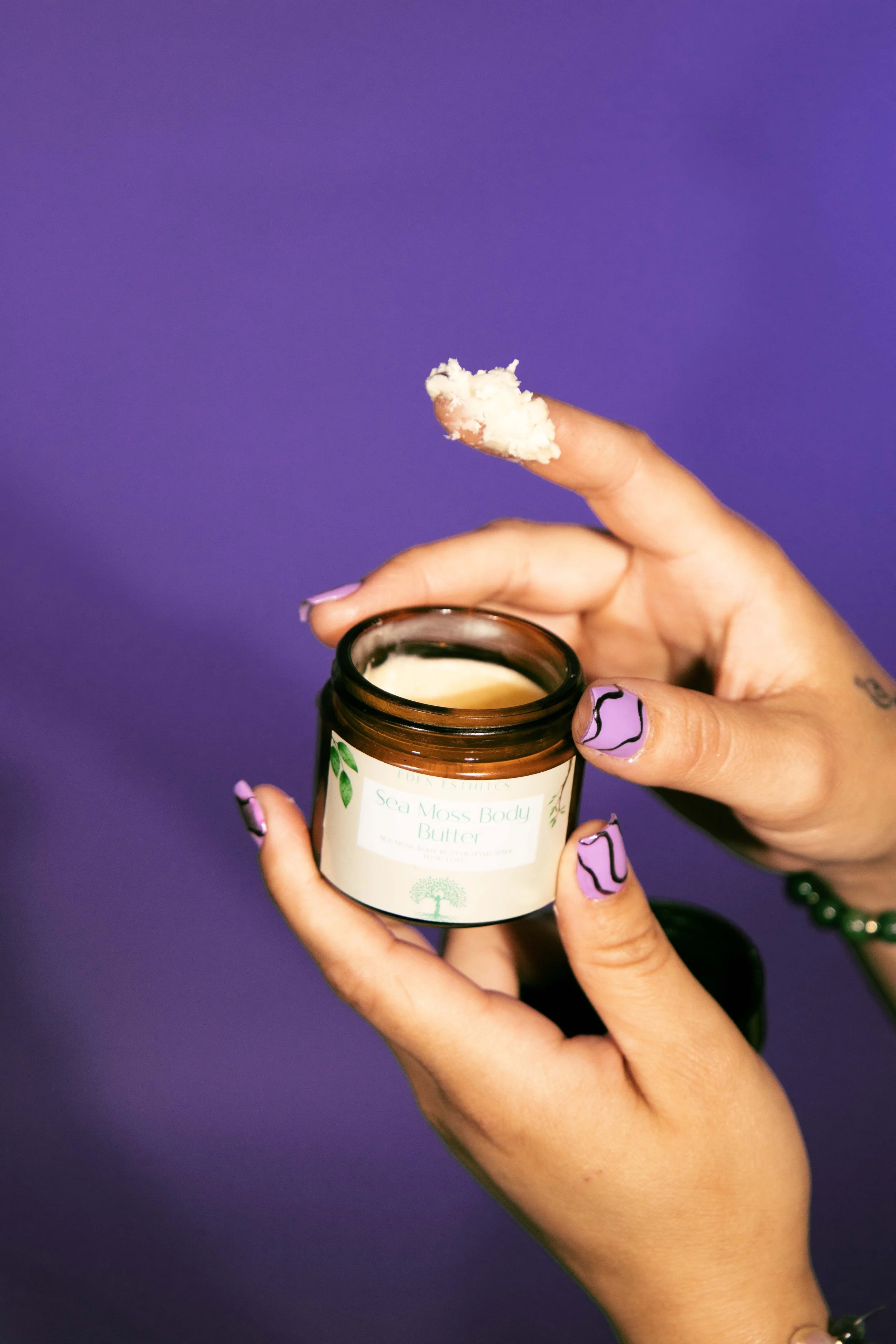 A person with purple painted nails holding a jar of sea moss body butter against a purple background.