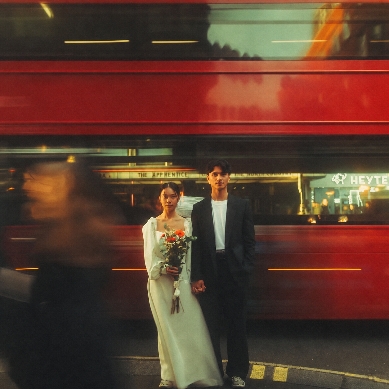 A young couple standing on a city street at night, with a moving red bus in the background. The woman is dressed in a white gown and holding a bouquet of flowers. The man is wearing a black suit and white shirt, standing next to her.