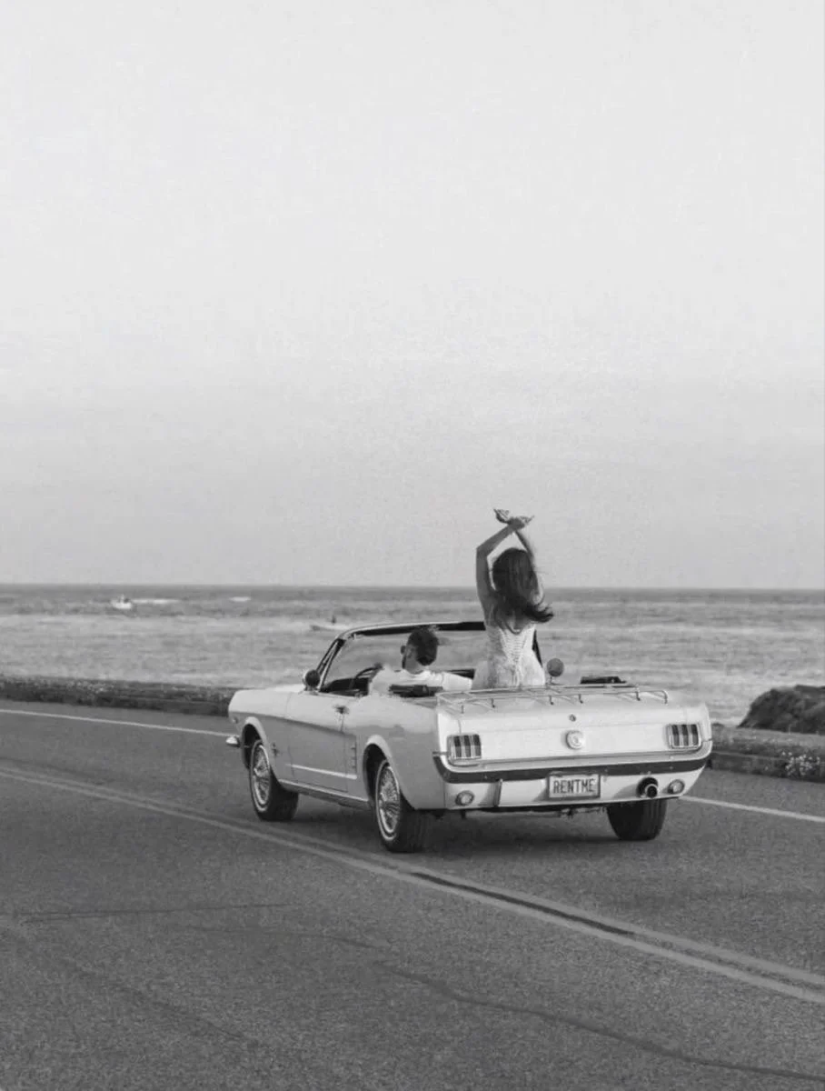 A black and white photo of a vintage convertible car driving along a seaside road with a woman sitting on the trunk holding a bird, and a man sitting in the driver's seat. The car has a "RENTME" license plate.