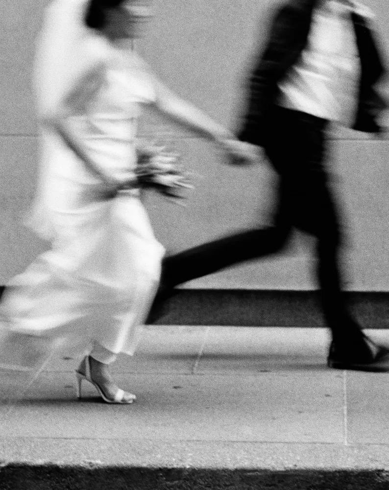 Blurry black-and-white photo of a woman in a white dress and high heels holding a bouquet, walking with a man in dark clothing and boots, with their motion captured in a rush.