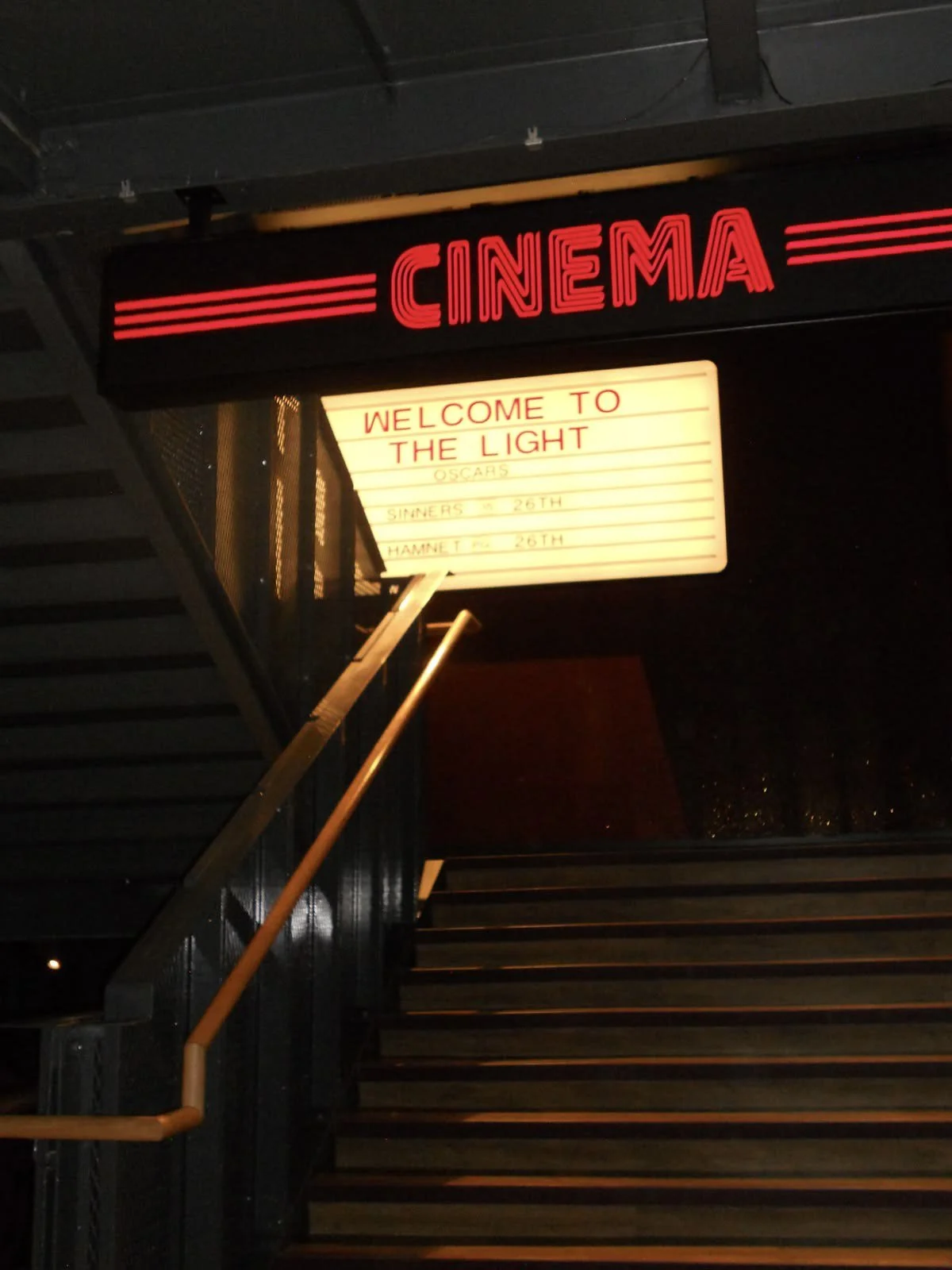 The image shows a cinema entrance with a neon sign labeled 'CINEMA' and an illuminated signboard displaying a welcome message for the movie 'The Light' along with other details.